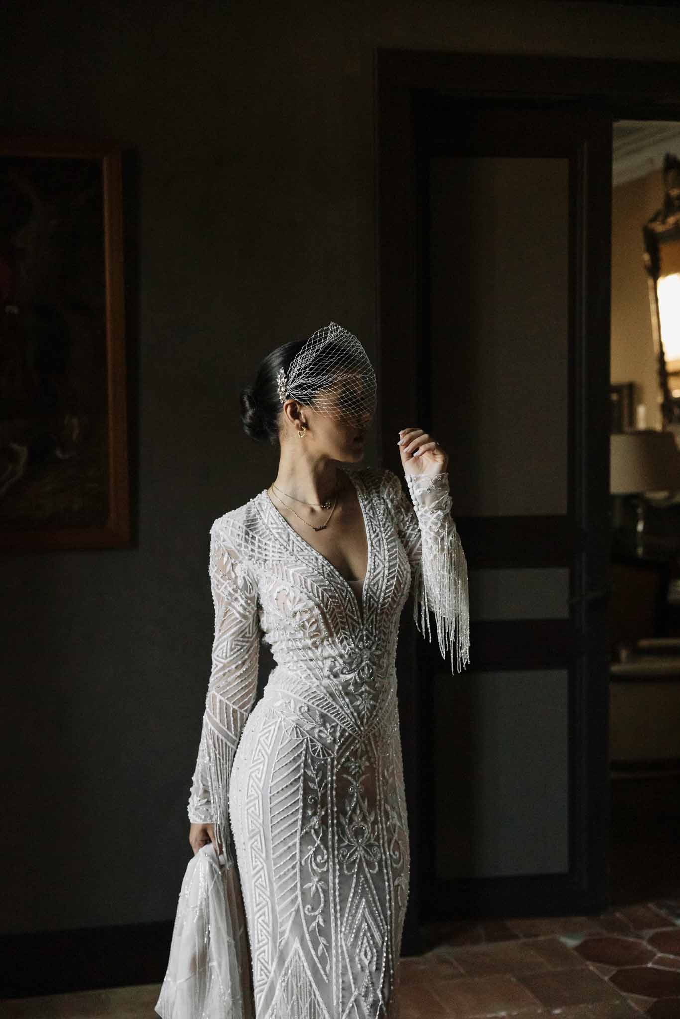 Bride in beaded Art Deco long-sleeve gown and birdcage veil standing in dimly lit chateau interior