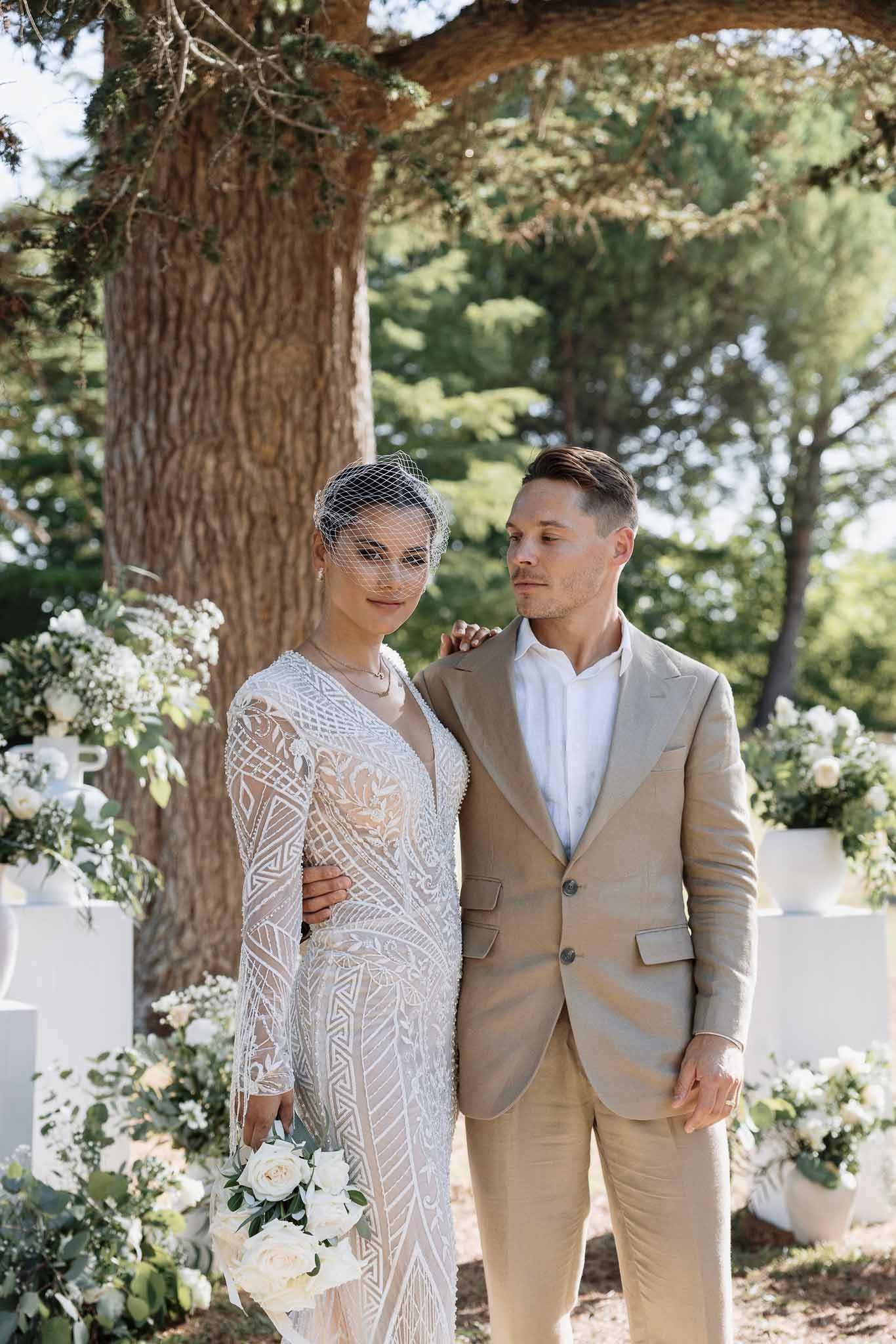 Bride in beaded long-sleeve gown with birdcage veil and groom in tan linen suit at outdoor ceremony backdrop