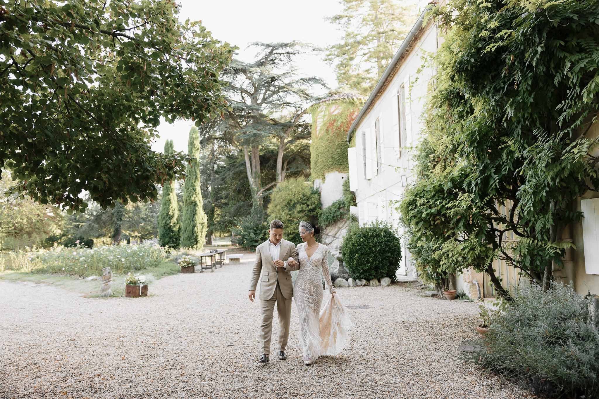 Couple walking arm in arm past ivy-covered country house, bride in beaded lace gown with birdcage veil