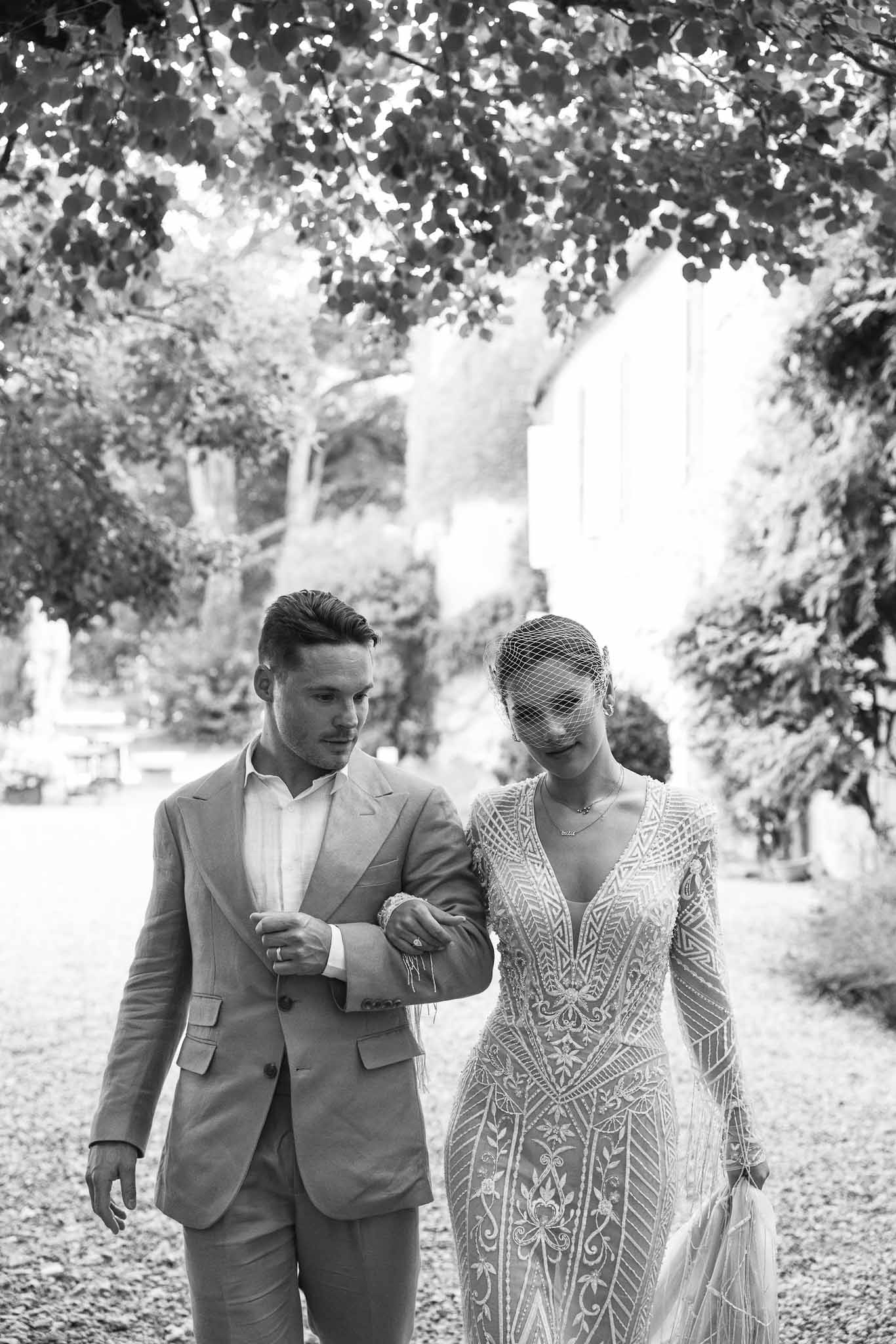 Black-and-white portrait of bride in beaded Art Deco gown and birdcage veil walking arm-in-arm with groom