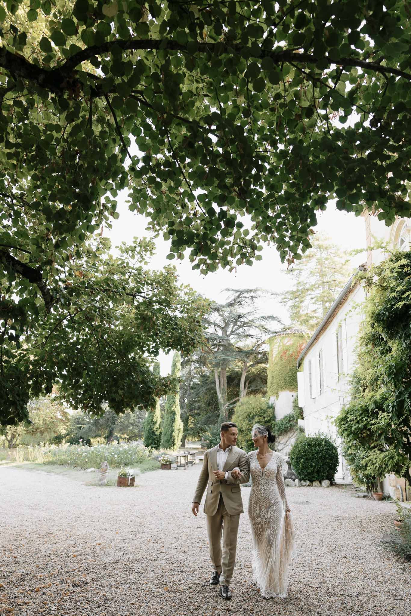 Couple walking arm-in-arm on gravel path, bride in beaded art deco gown with birdcage veil, before ivy-covered manor