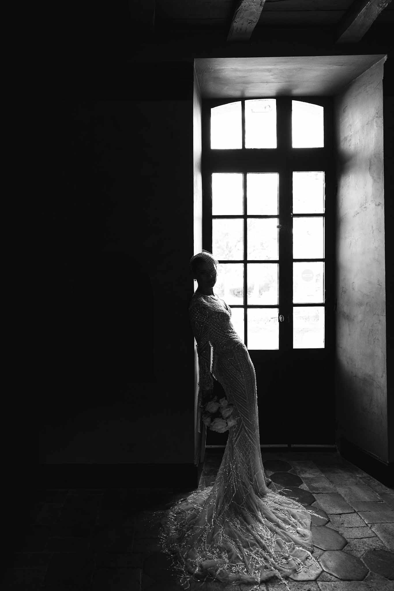 Black and white portrait of bride in beaded long-sleeve gown silhouetted against arched window