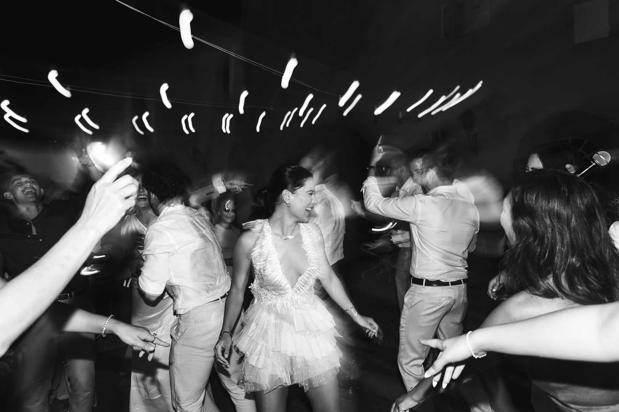 Black and white motion-blur bride in ruffle mini dress dancing with guests under streaking bulb lights
