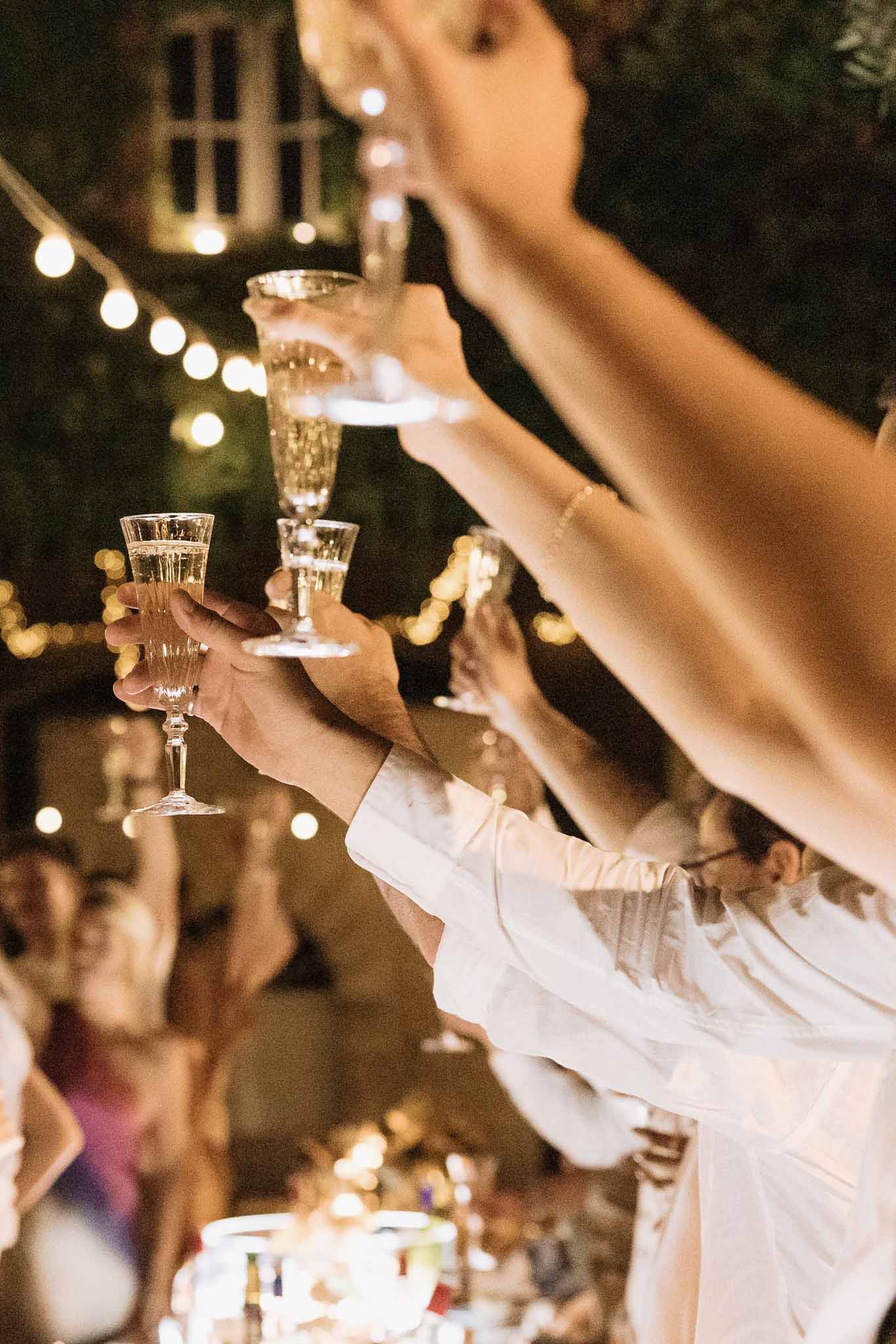 Guests raising champagne flutes in toast during evening outdoor reception with string lights overhead