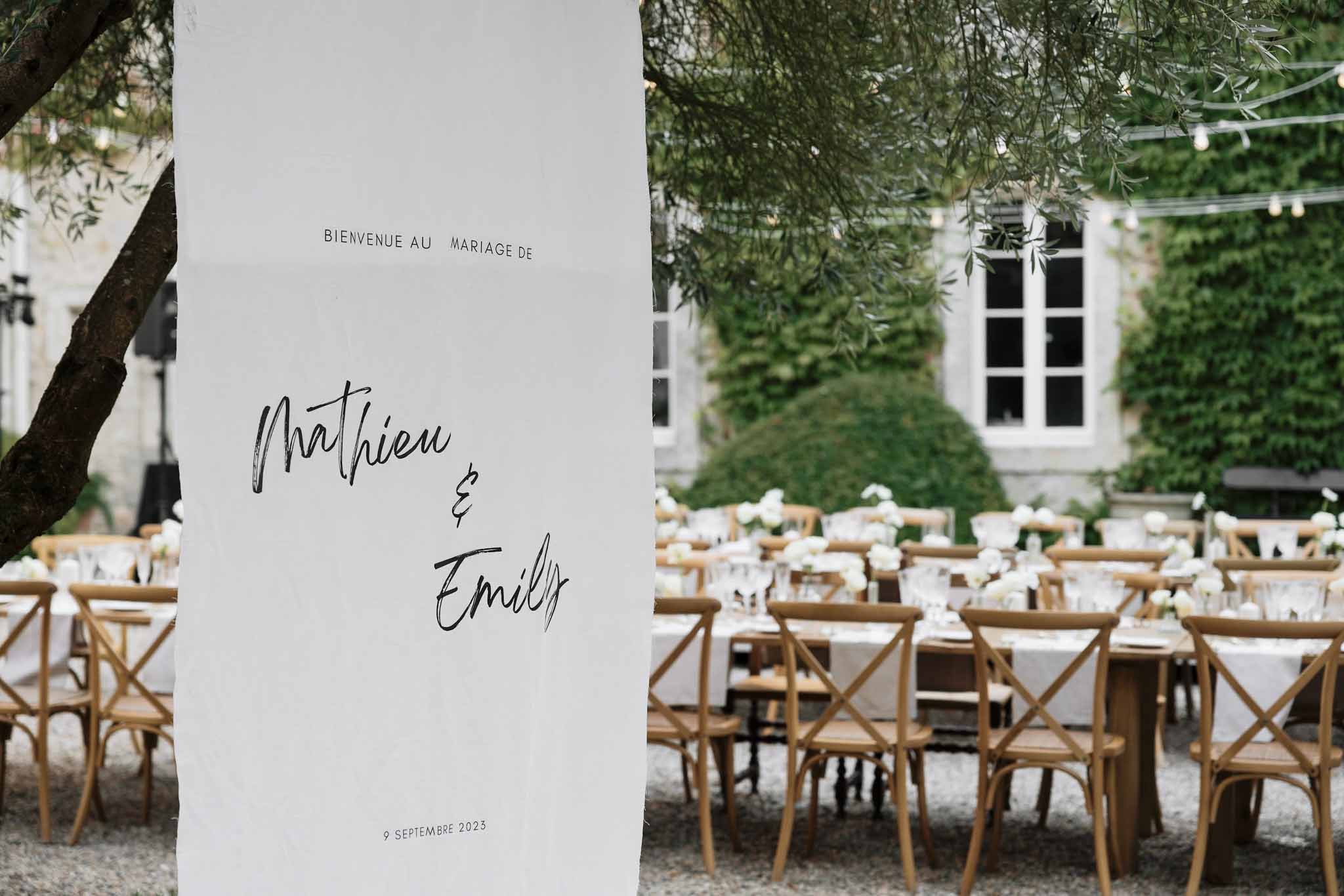 Courtyard reception with Bienvenue sign fairy lights white rose centerpieces and cross-back chairs