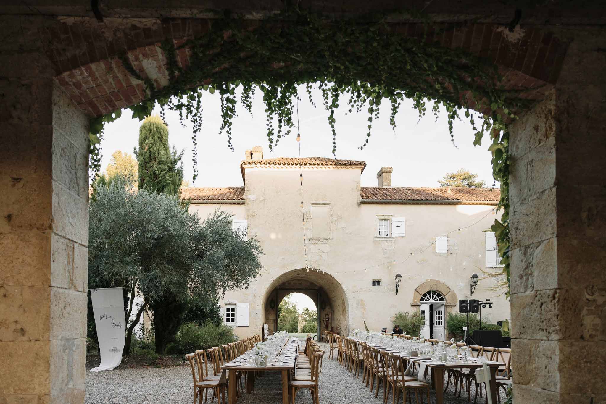 Farm tables through ivy-draped stone archway with calligraphy banner and festoon lights in cream-rendered courtyard