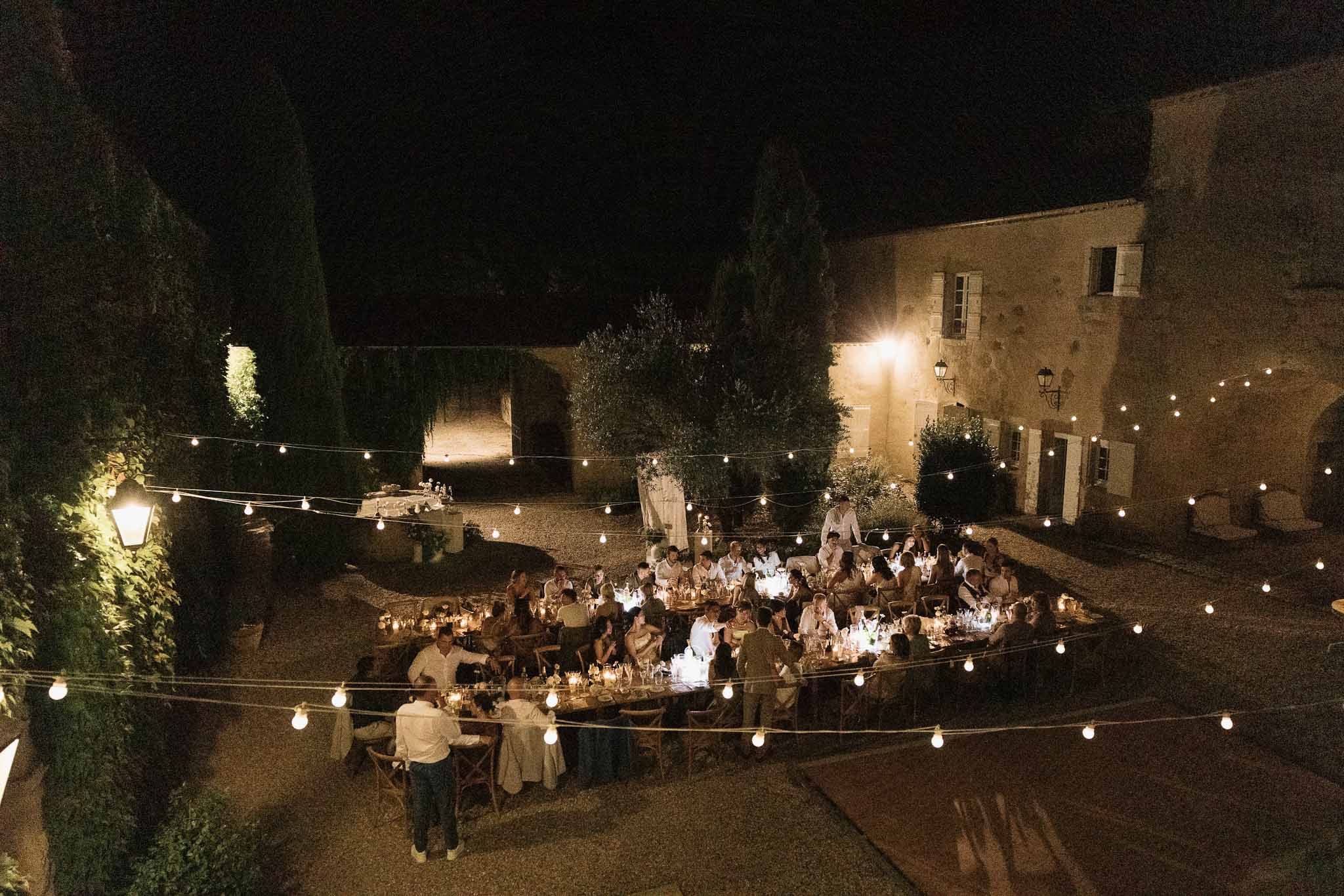 Elevated view of 70 guests dining under festoon lights in stone courtyard at French mas at night