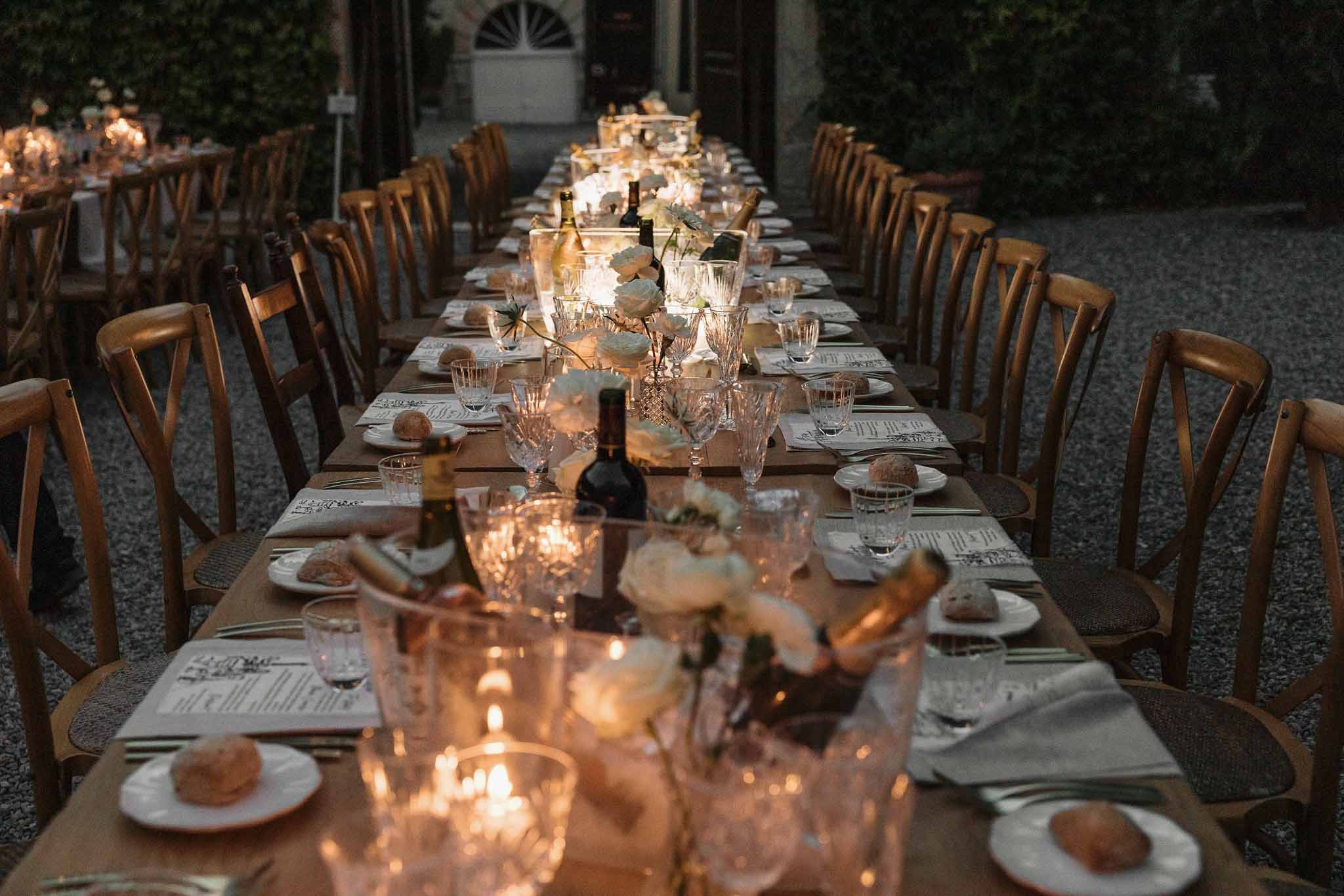 Long banquet table at dusk with white roses, votive candles, crystal glassware, and printed menus on a gravel courtyard