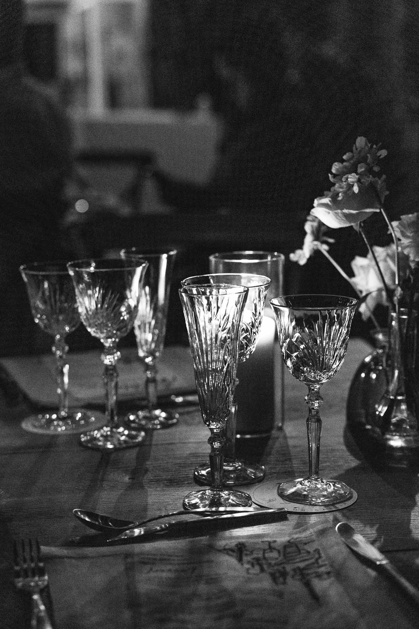 Crystal glasses and silverware on wooden reception table in candlelight captured in moody black and white