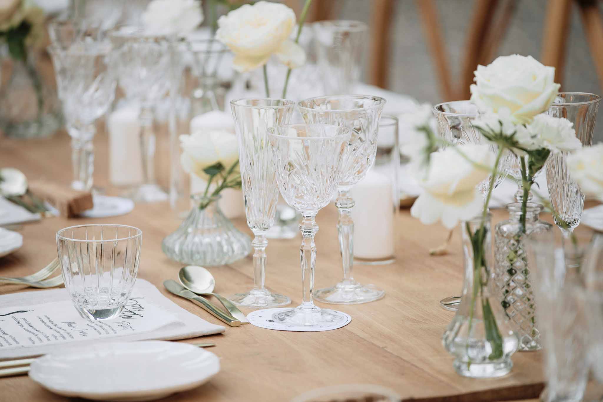 Reception table detail with crystal glasses, ivory roses, white pillar candles, gold flatware on wooden farm table