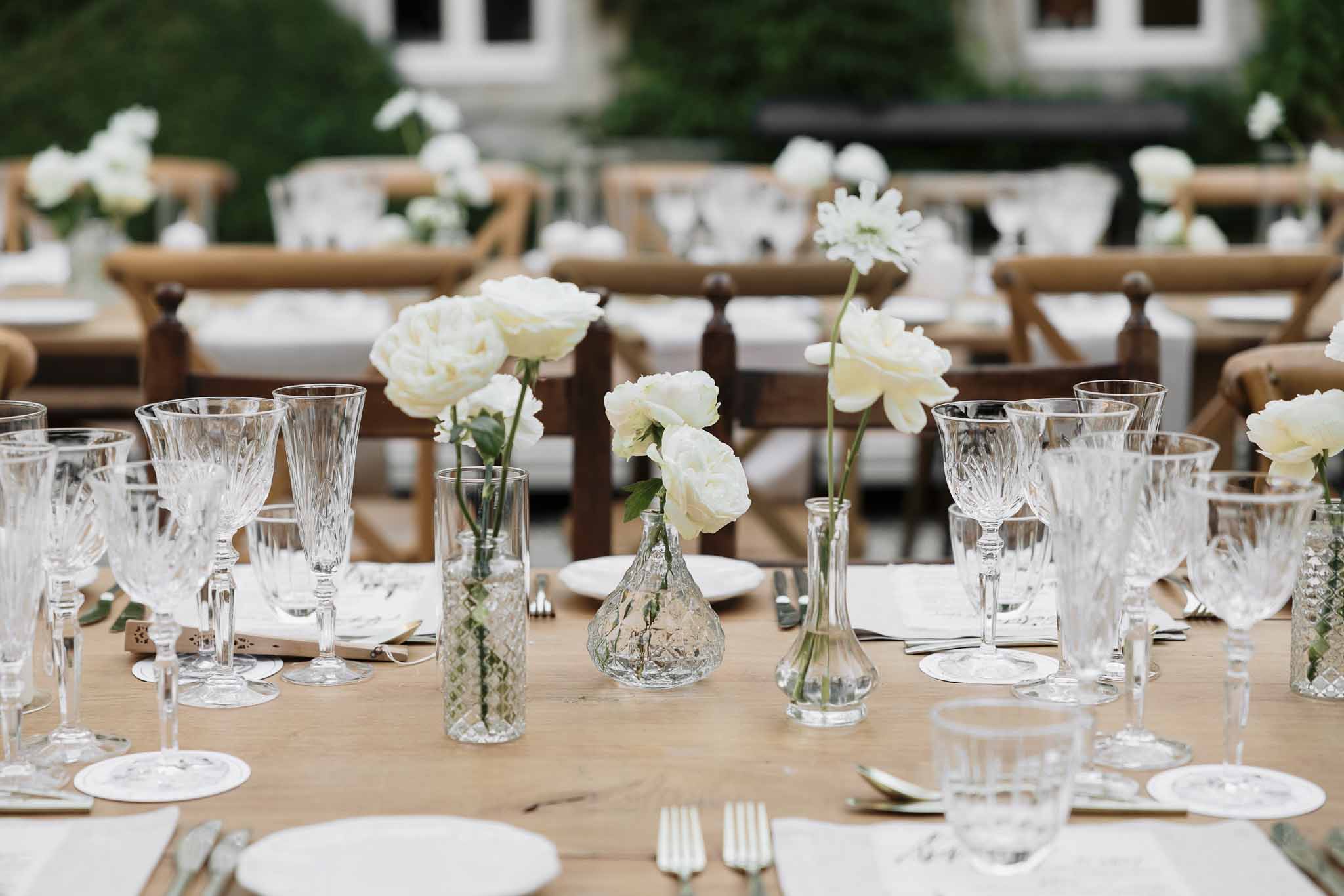 Farm table with assorted glass bud vases holding ivory roses ranunculus and scabiosa cross-back chairs and chateau behind