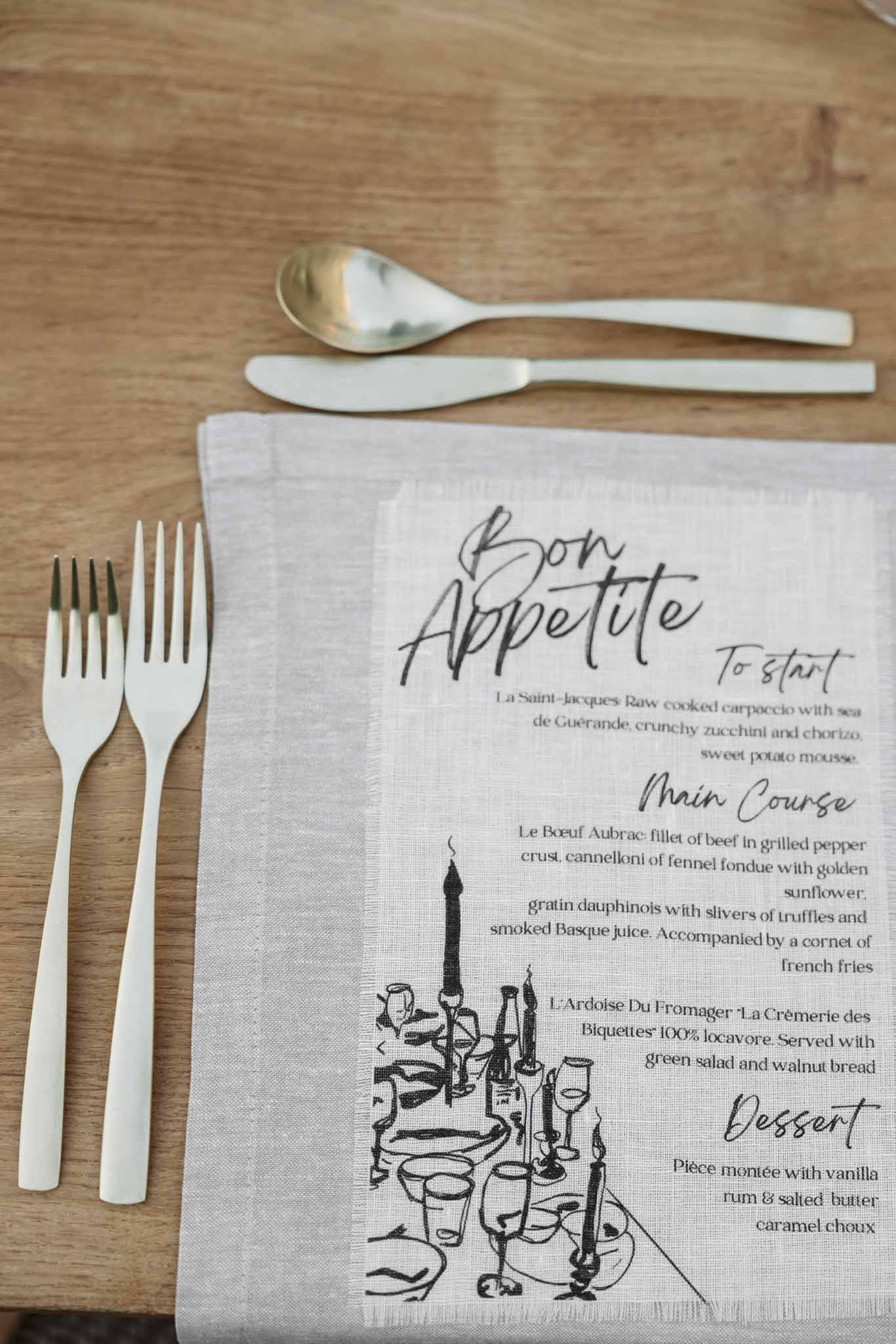 Flat lay of wedding menu printed on grey linen napkin with matte white cutlery and gold spoon on natural wood table