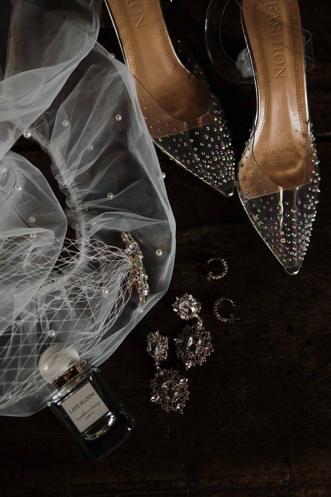 Crystal PVC heels, pearl-accent veil, gold statement earrings, and perfume on dark wood surface