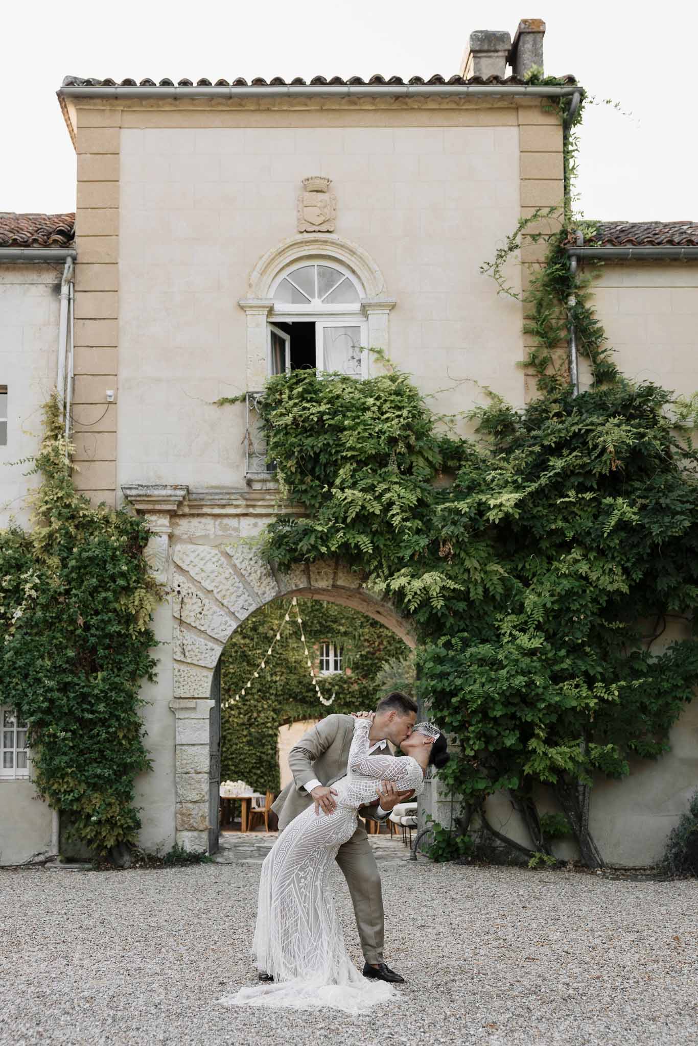 Groom dipping bride for a kiss under vine-covered stone archway of French chateau on gravel courtyard