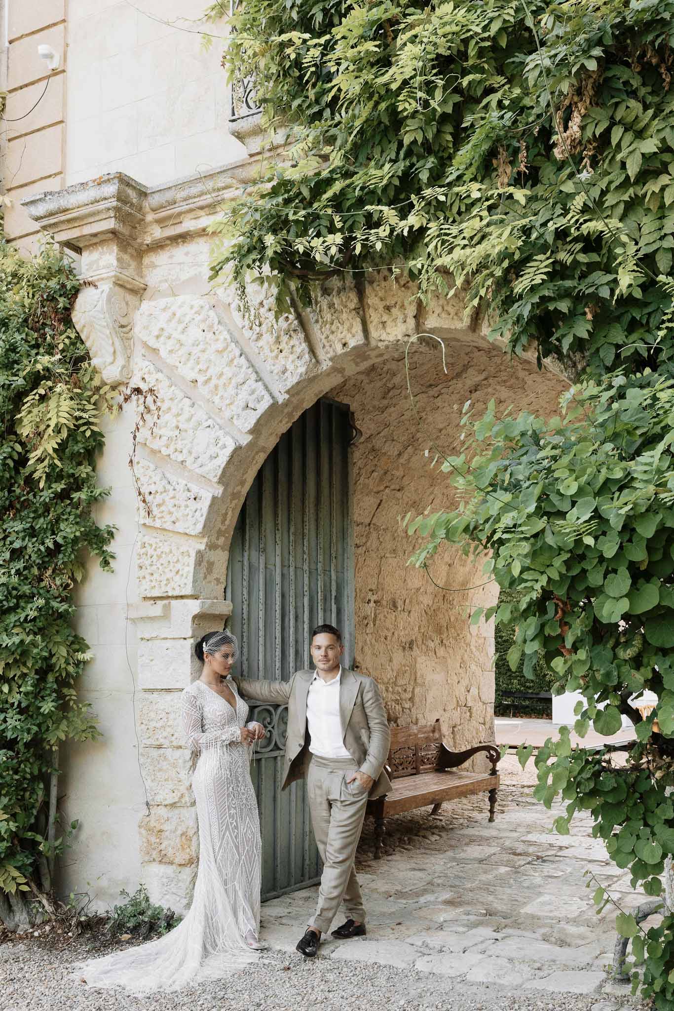 Bride in geometric beaded gown with birdcage veil and groom in taupe suit at arched chateau door
