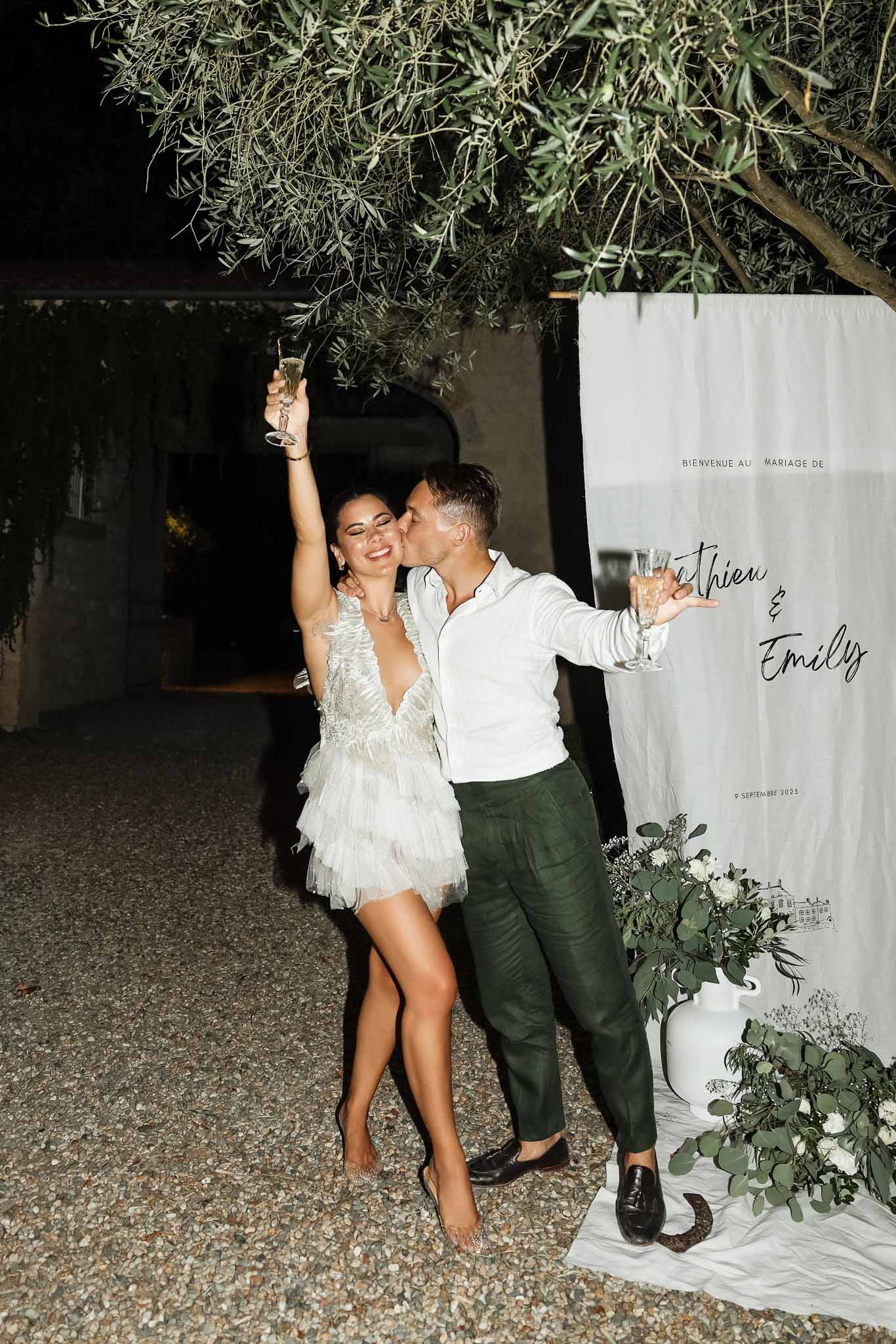 Couple with champagne at night beside bienvenue sign with eucalyptus, bride in feathered mini dress