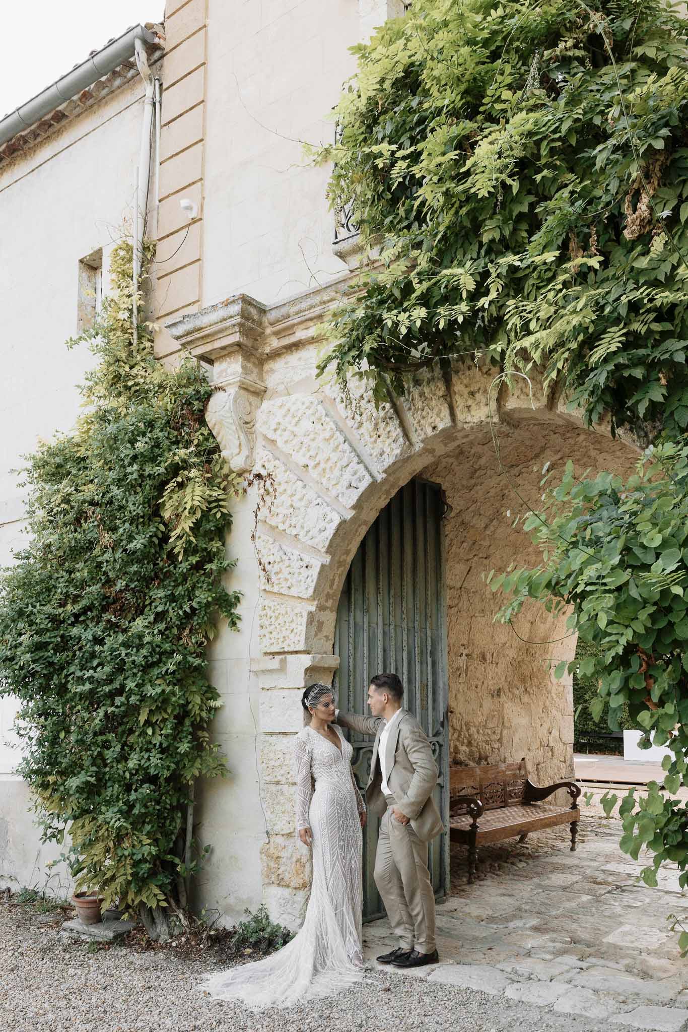 Bride and groom standing beneath ornate stone archway with green wooden door at French chateau