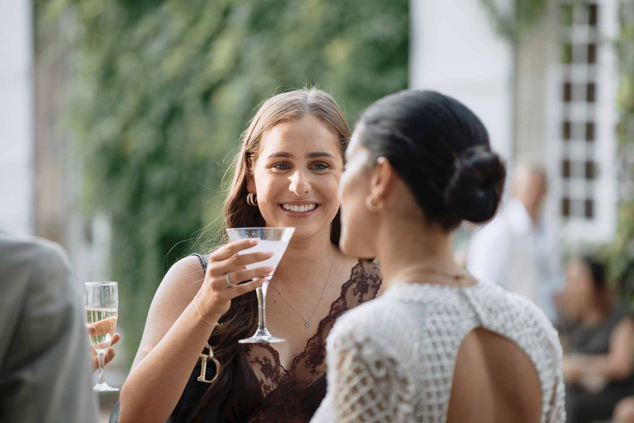 Wedding guest in navy lace dress holding cocktail glass chatting with bride at outdoor cocktail hour