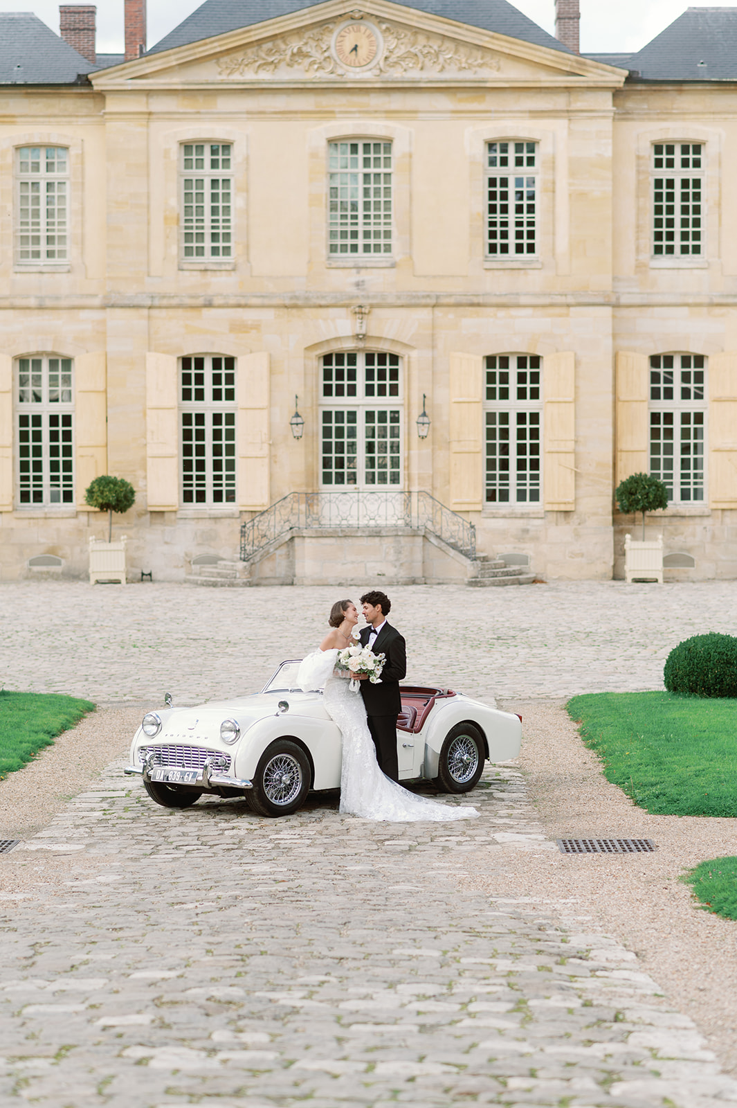 Bride and groom beside vintage white Triumph convertible in front of classical French chateau facade