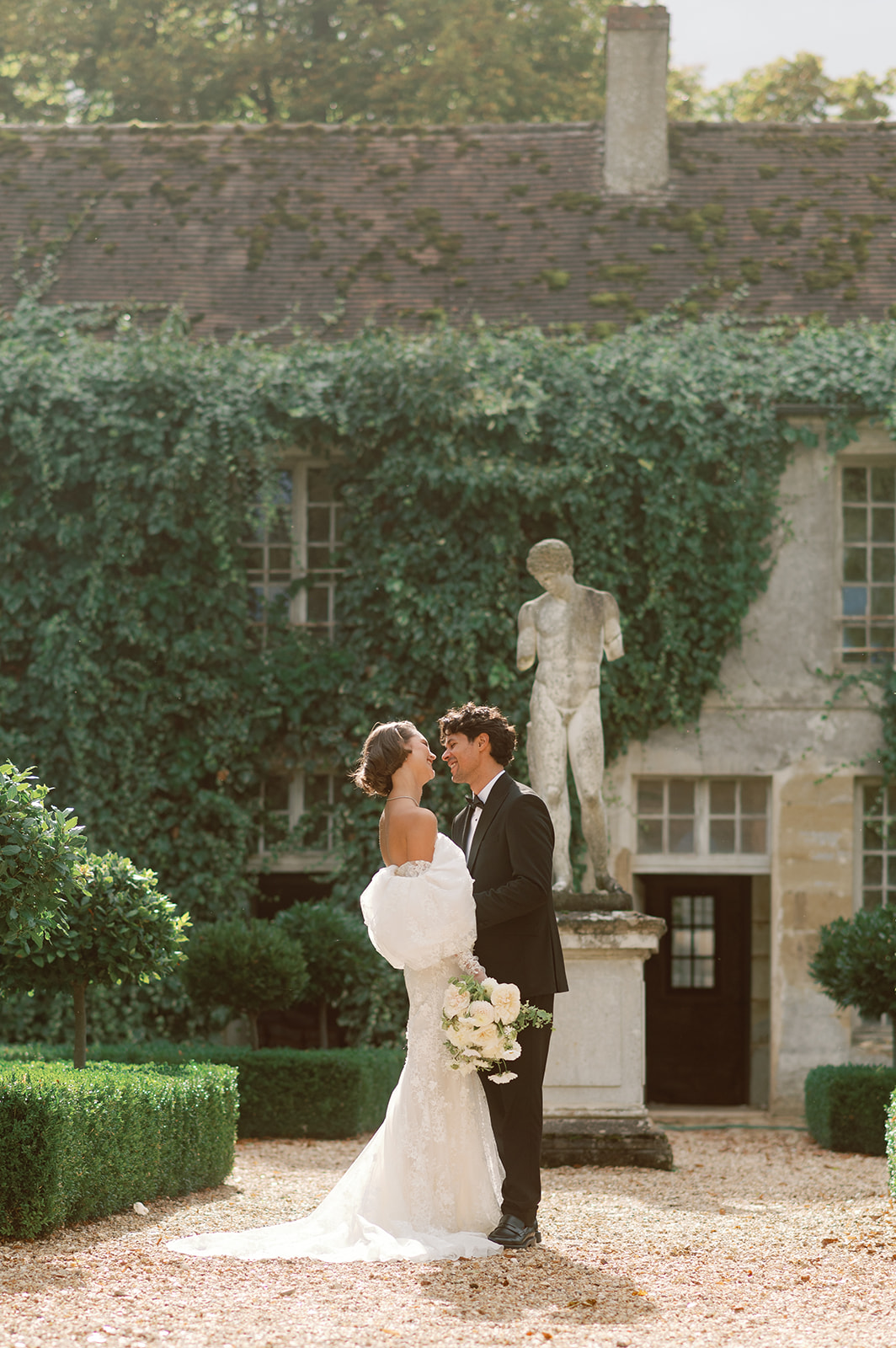Bride in ivory lace gown with fur stole and groom in black tuxedo smiling in chateau formal garden
