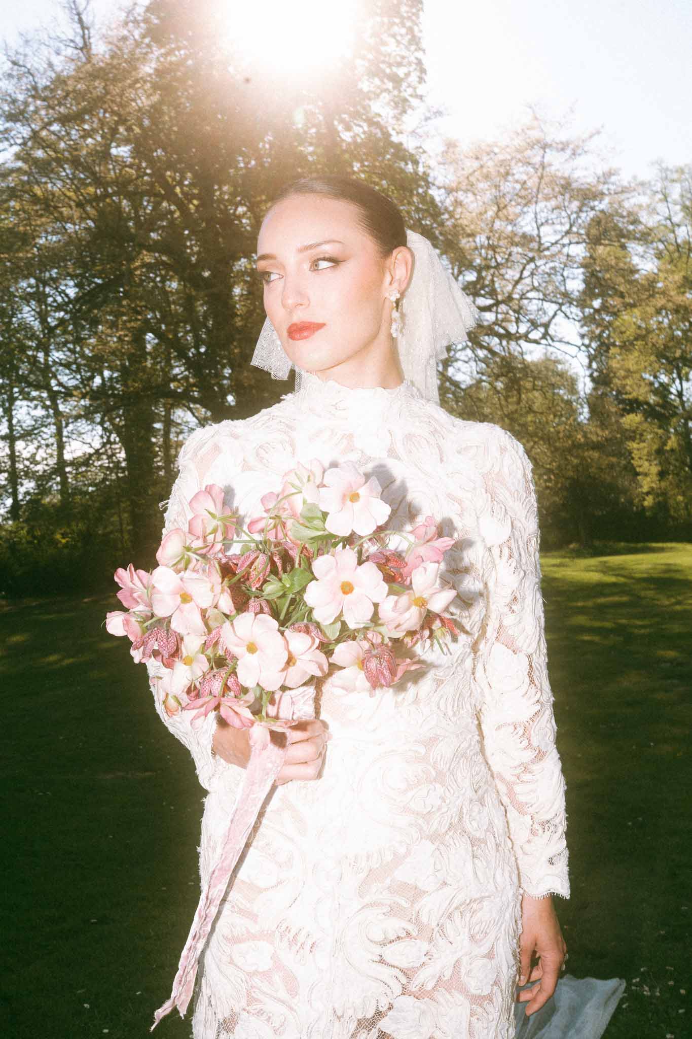 Bride in ivory lace dress holding white and pink bouquet in tree-lined park setting