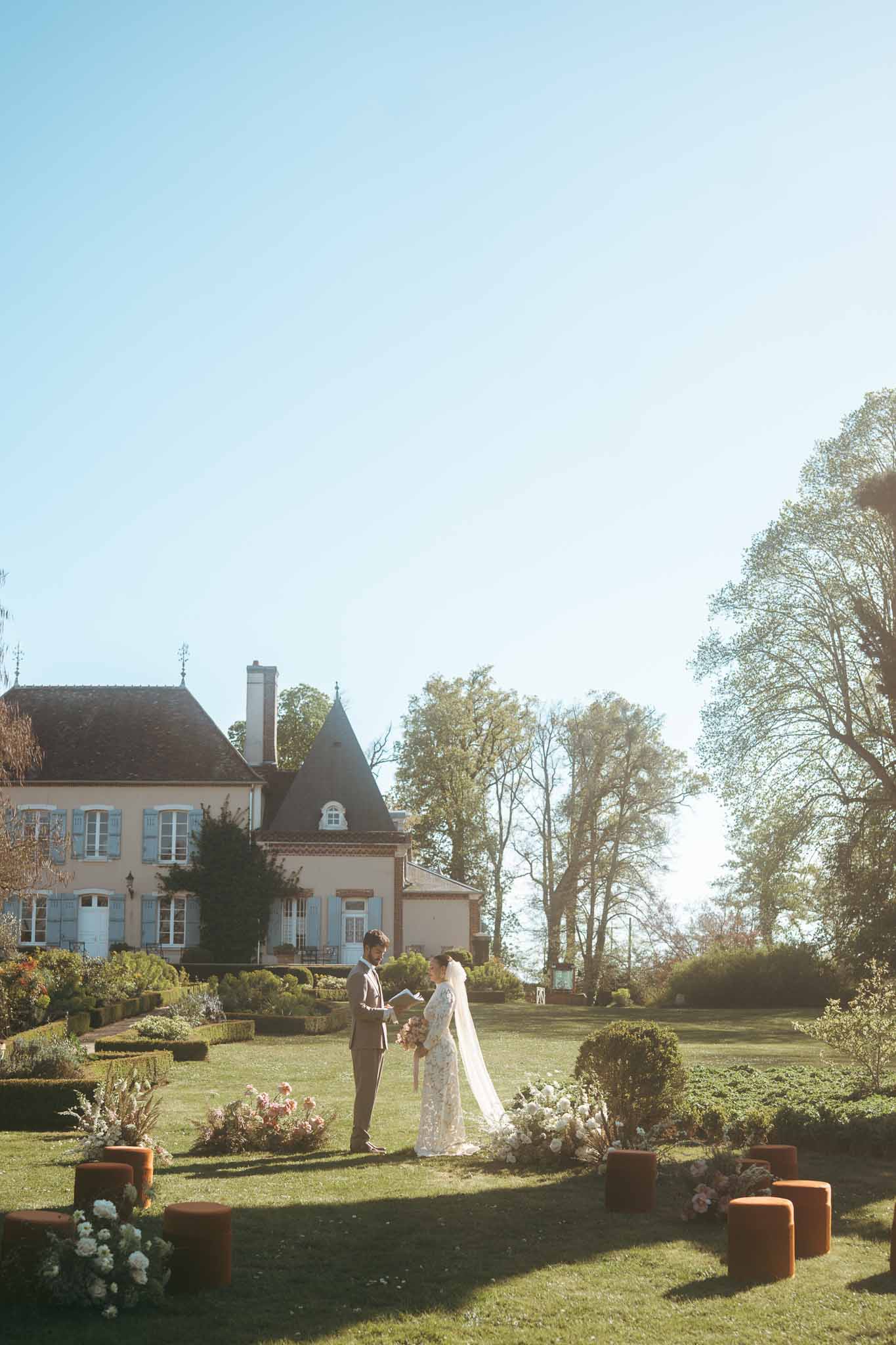 Bride and groom ceremony in manicured gardens of French manor house with formal flower arrangements