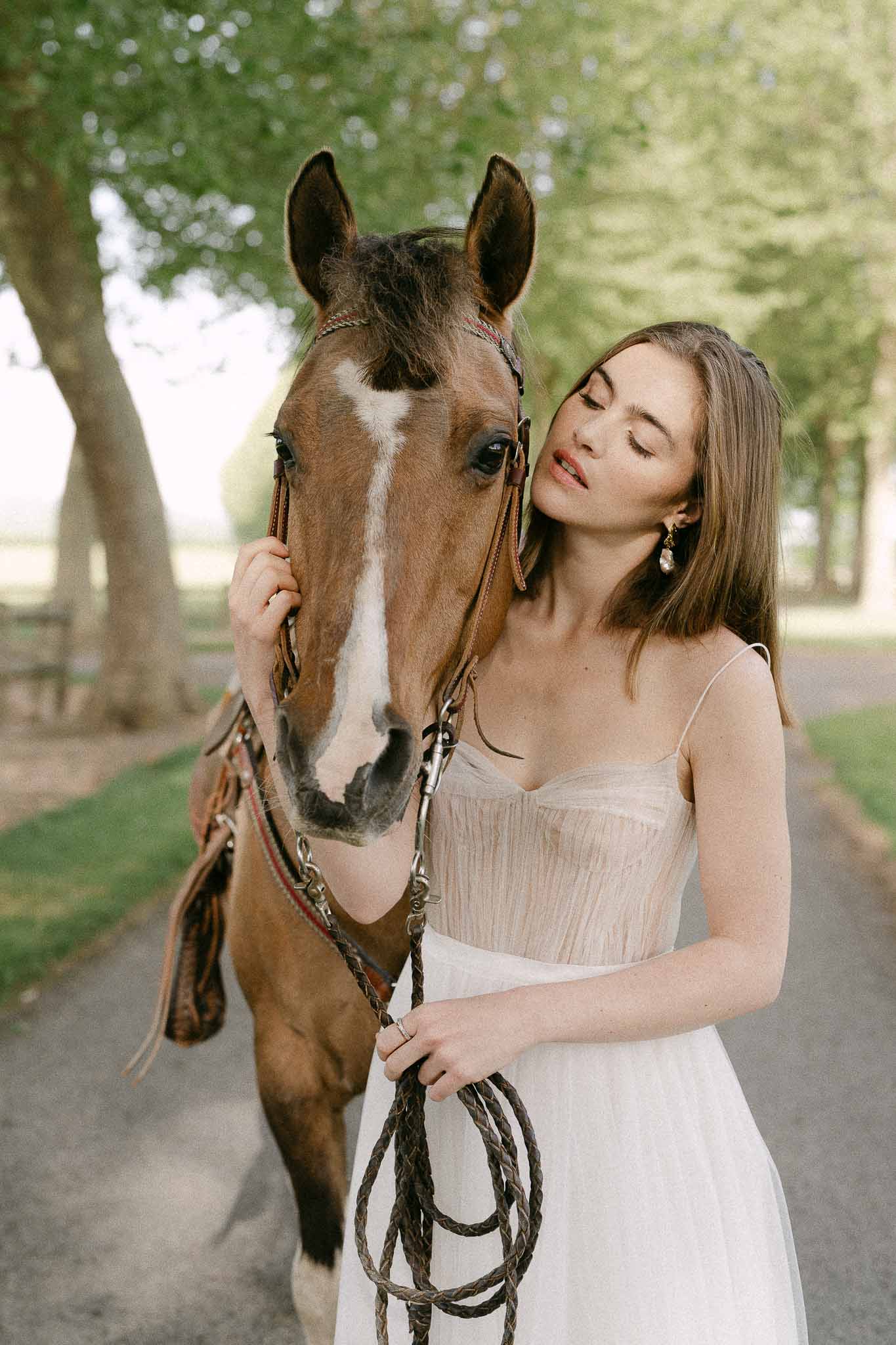 Bride in cream dress posing with chestnut horse in tree-lined countryside setting