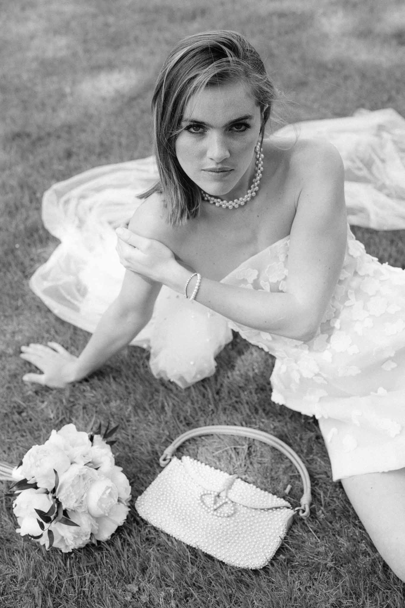 Black and white bridal portrait of bride in off-shoulder dress with pearl jewelry seated on grass outdoors