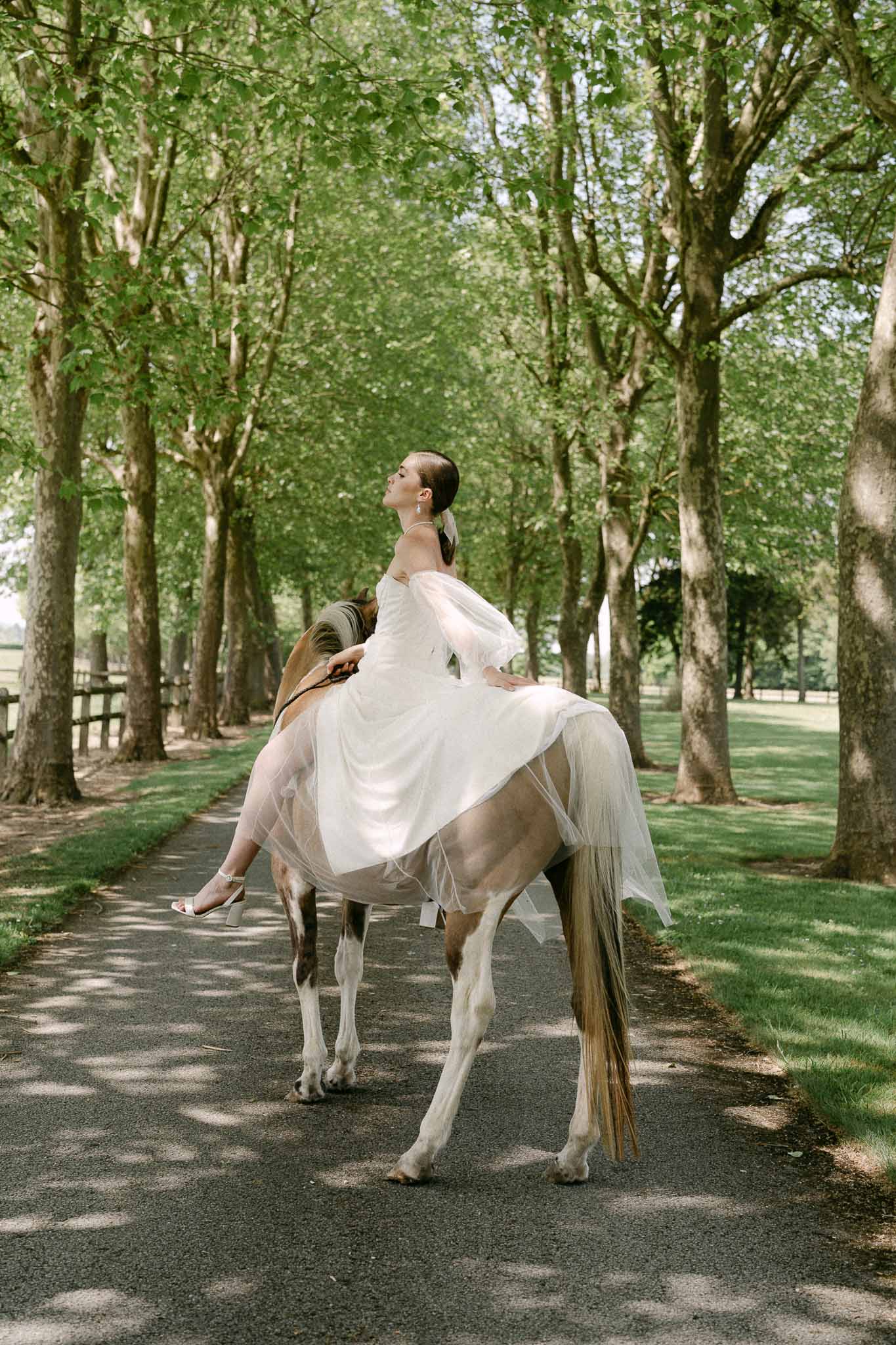 Bride on horseback riding down tree-lined avenue at outdoor estate wedding venue