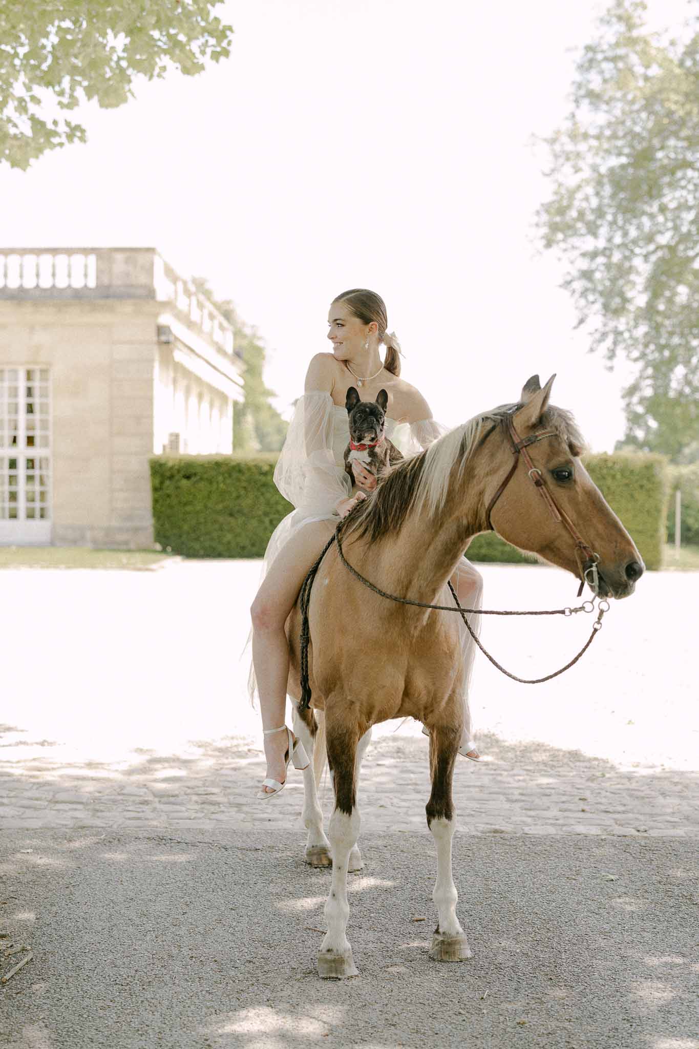 Bride on horseback holding French Bulldog in classical chateau courtyard