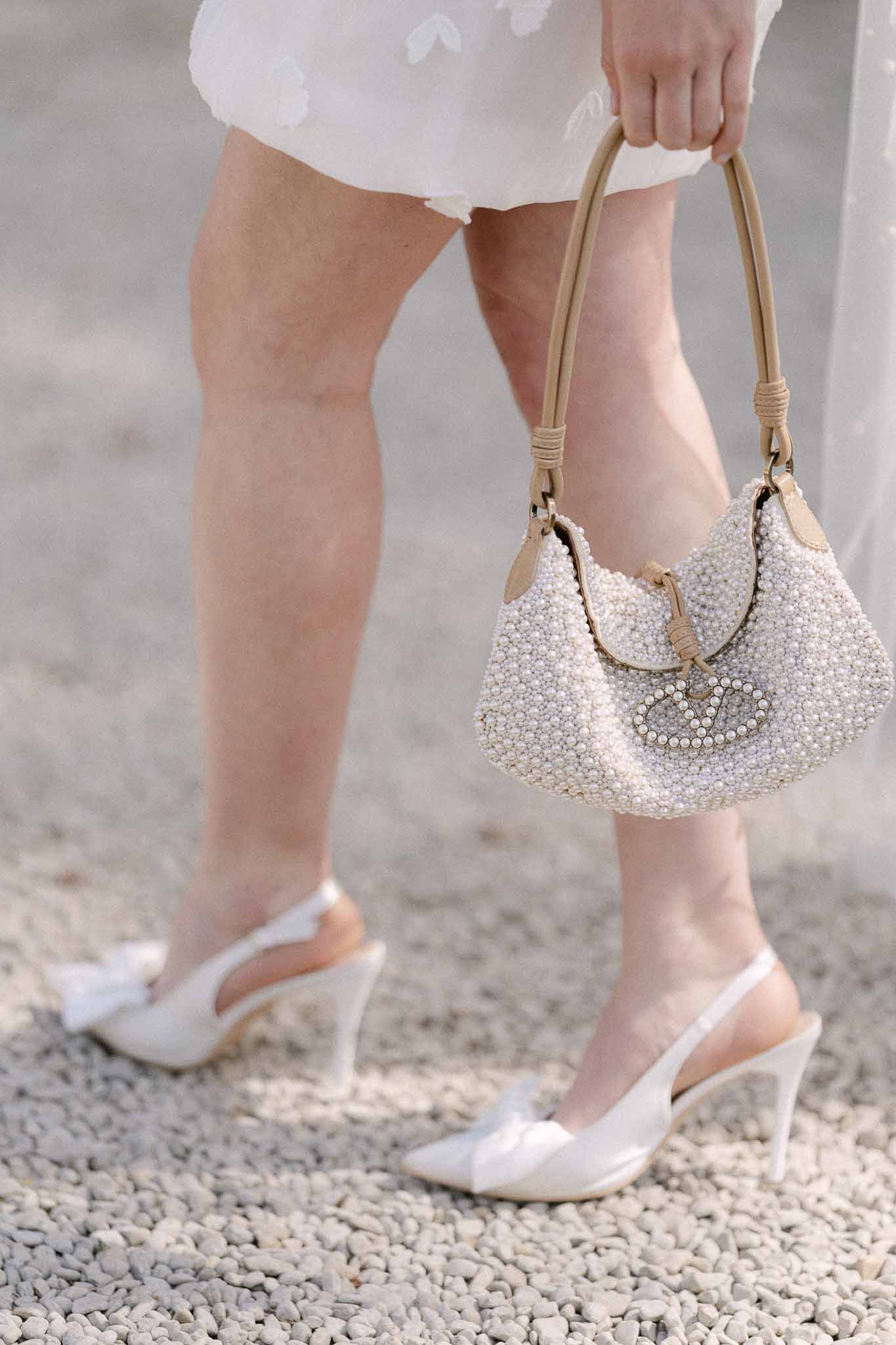 Bride's ivory dress, satin heels and pearl bag on gravel pathway at wedding venue