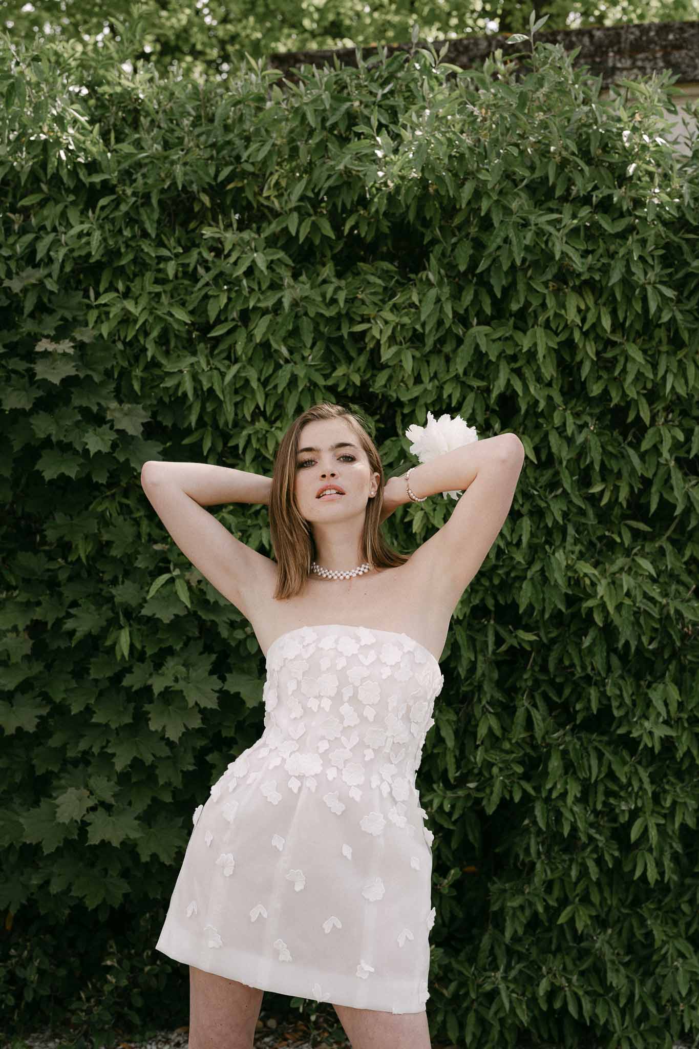 Bride in white petal appliqué dress posing against lush green foliage backdrop
