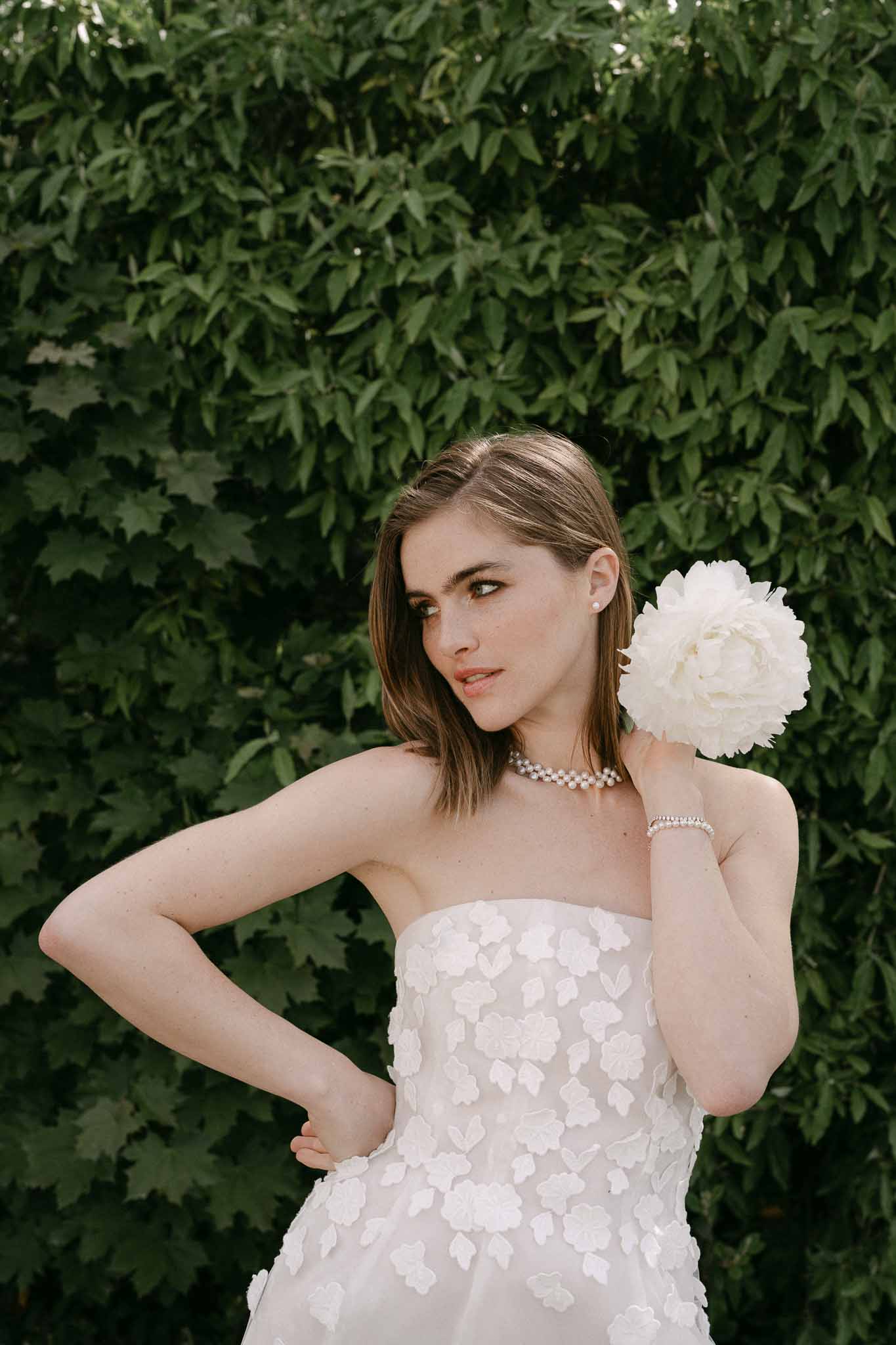 Bride in floral appliqué wedding dress holding peony bouquet against ivy wall
