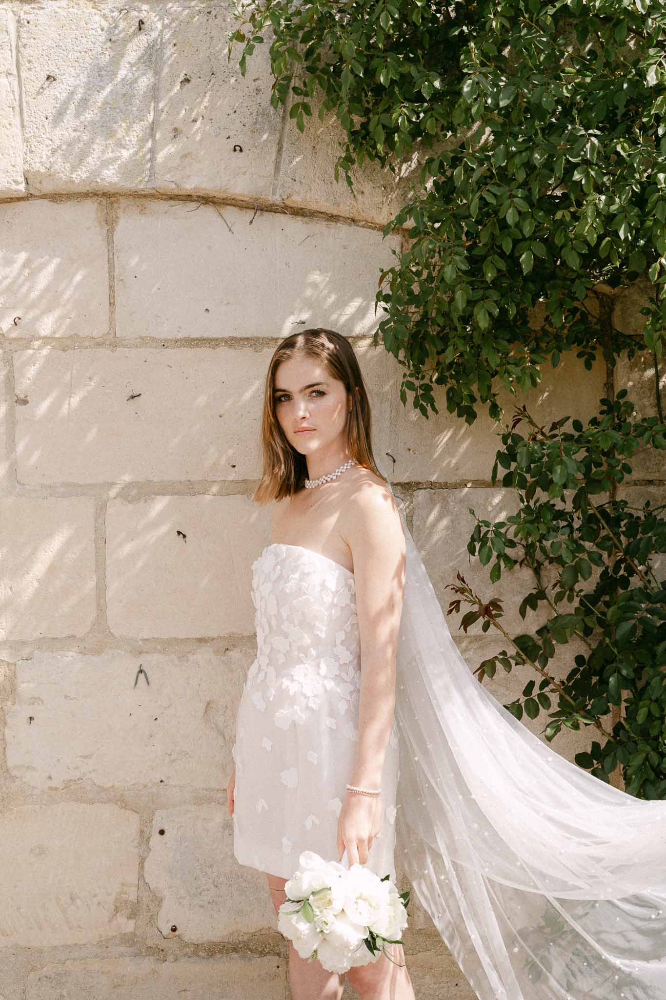 Bride in strapless white dress with veil against stone wall with ivy