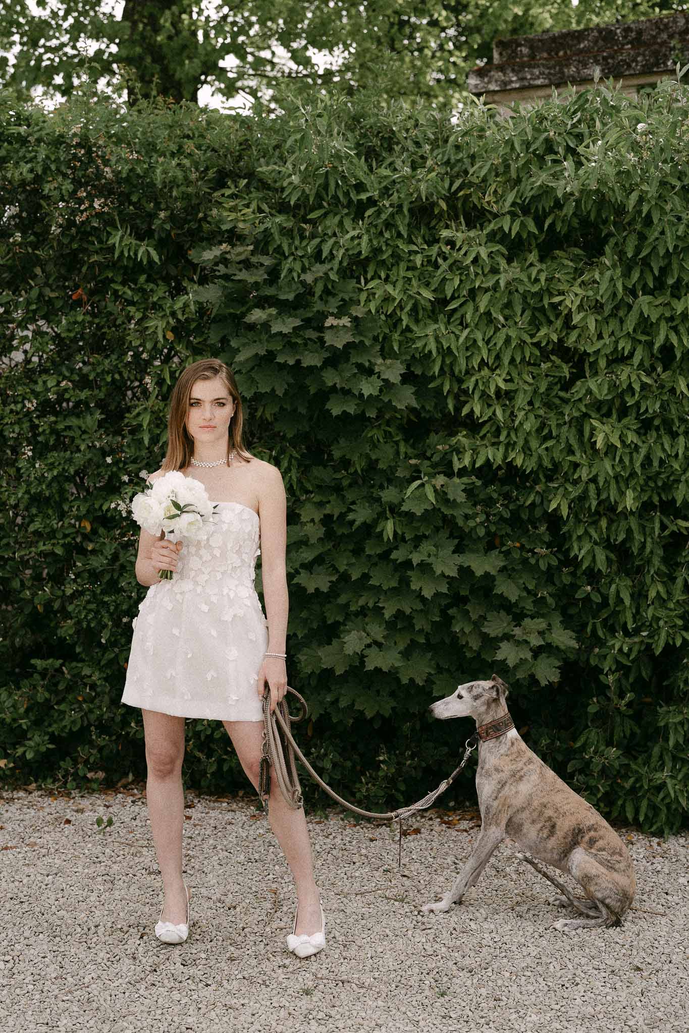Bride in white dress with bouquet and greyhound dog on gravel pathway at ivy-covered stone venue