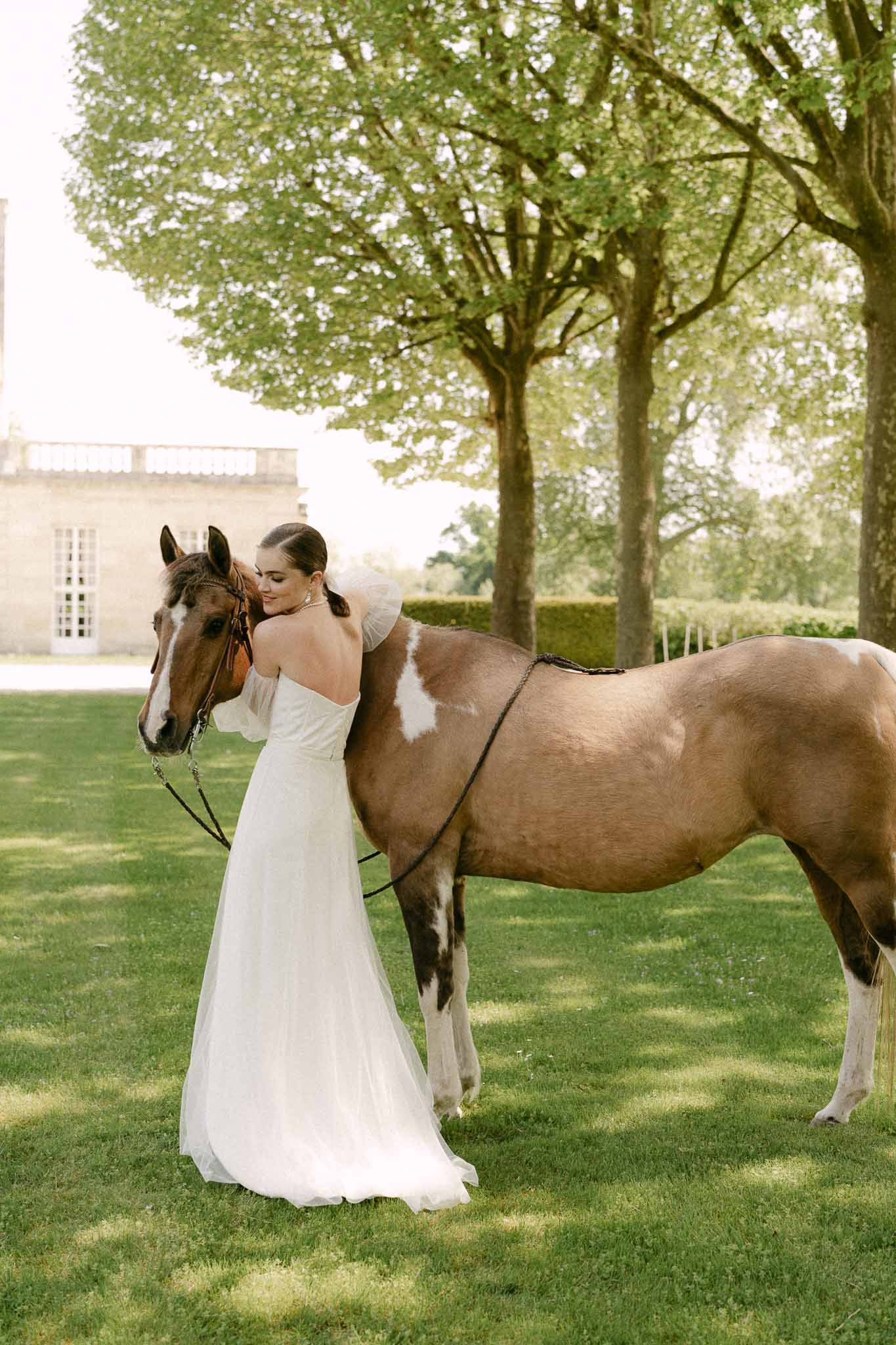 Bride in ivory gown posing with brown horse on estate grounds with linden trees