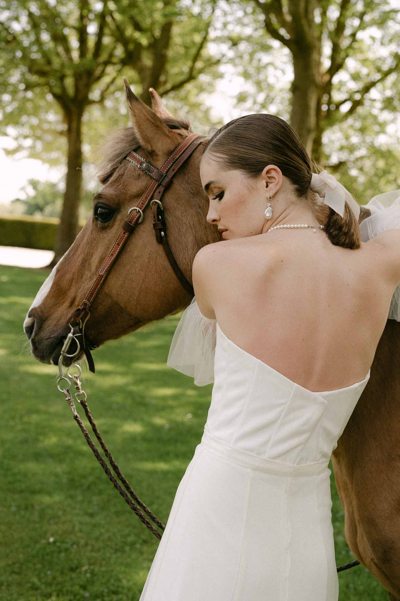 Bride in ivory strapless dress posing with chestnut horse in tree-lined garden setting