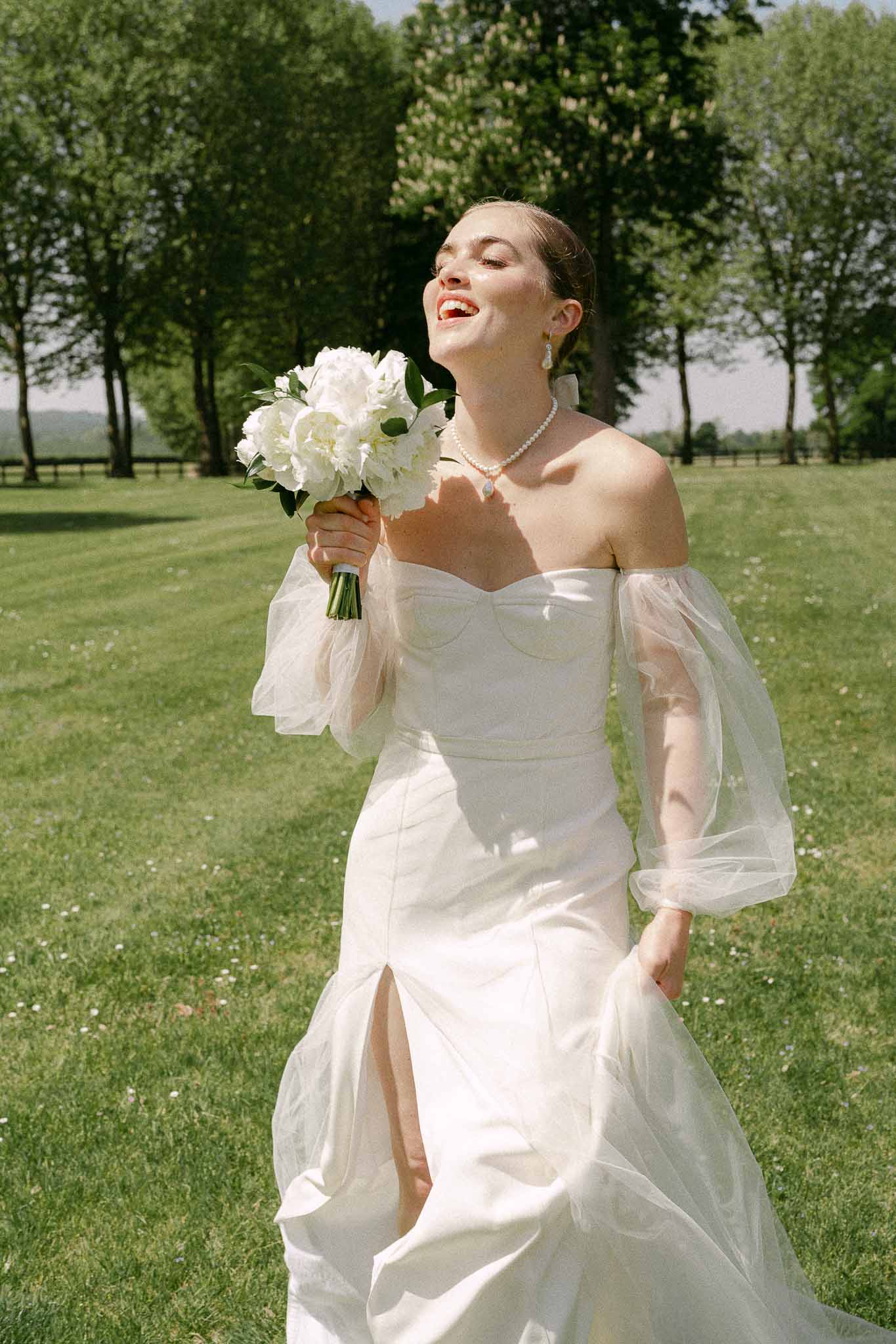 Bride in ivory mermaid gown holding peony bouquet in garden setting with tree-lined background