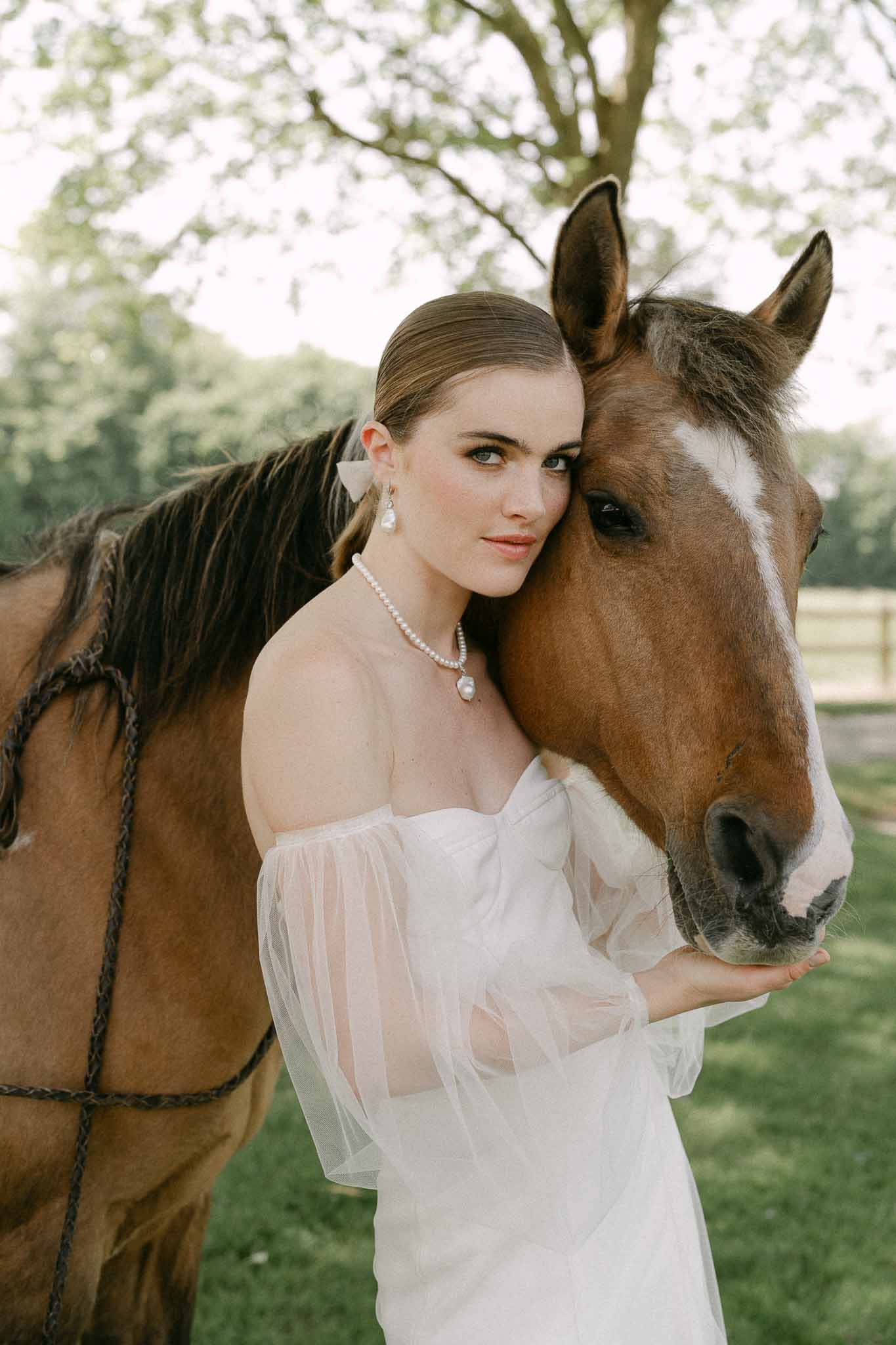 Bride in ivory off-shoulder dress poses with chestnut horse at countryside estate