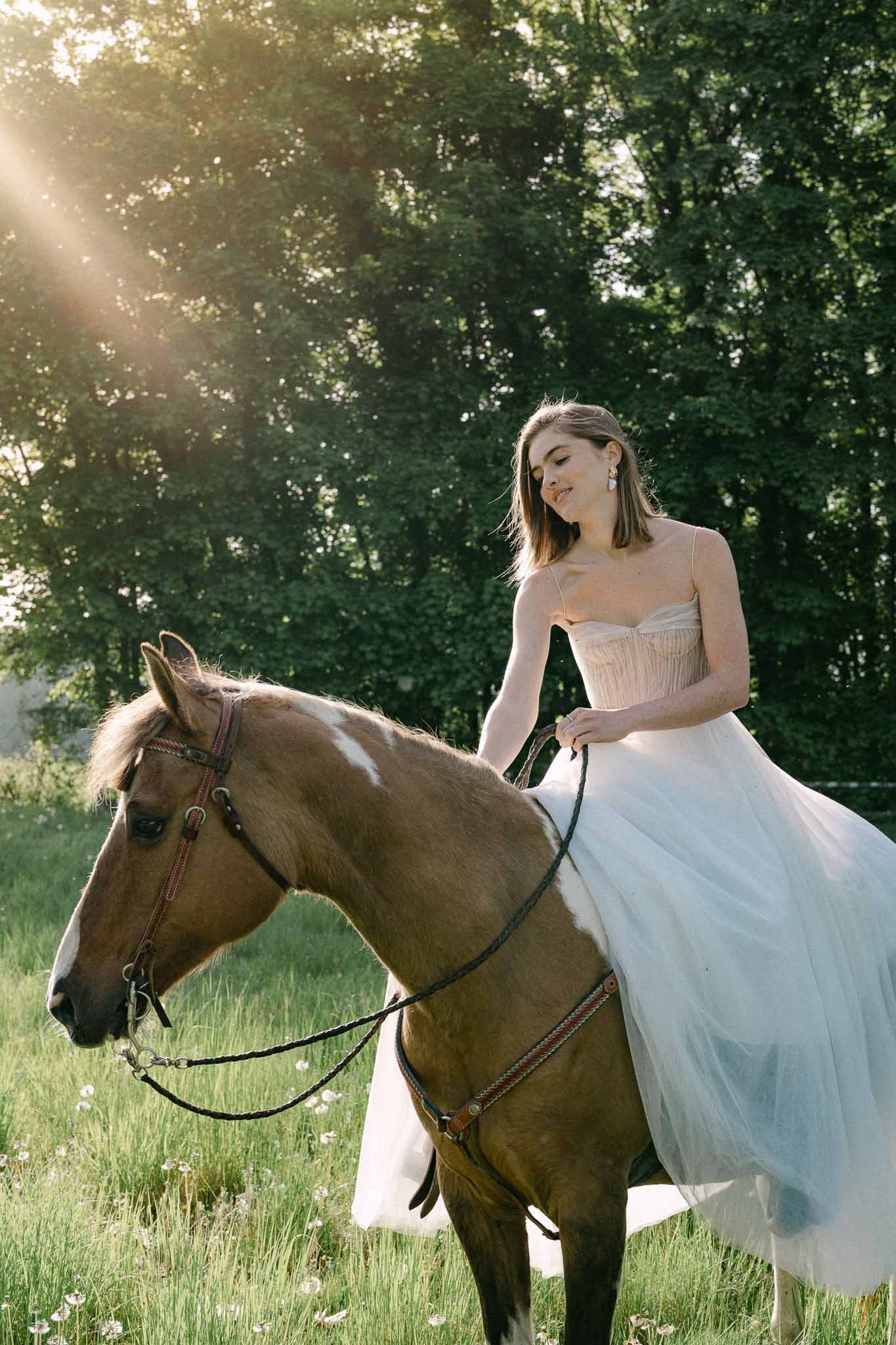 Bride in ivory gown riding chestnut horse in countryside meadow with trees