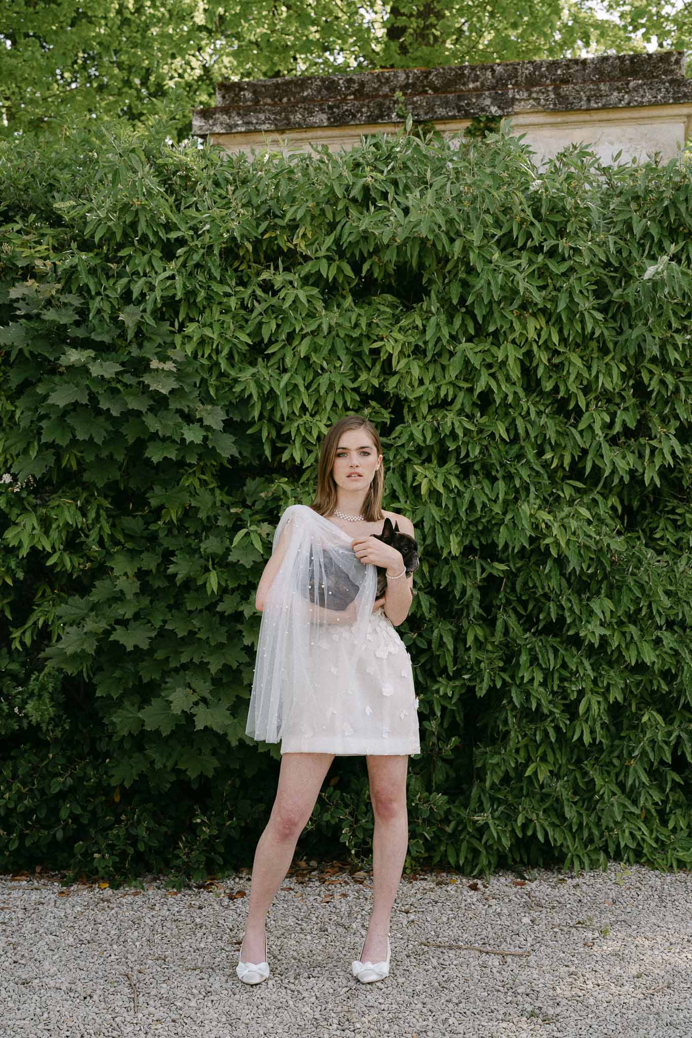 Bride in ivory dress holding black cat in garden setting with ivy-covered stone wall