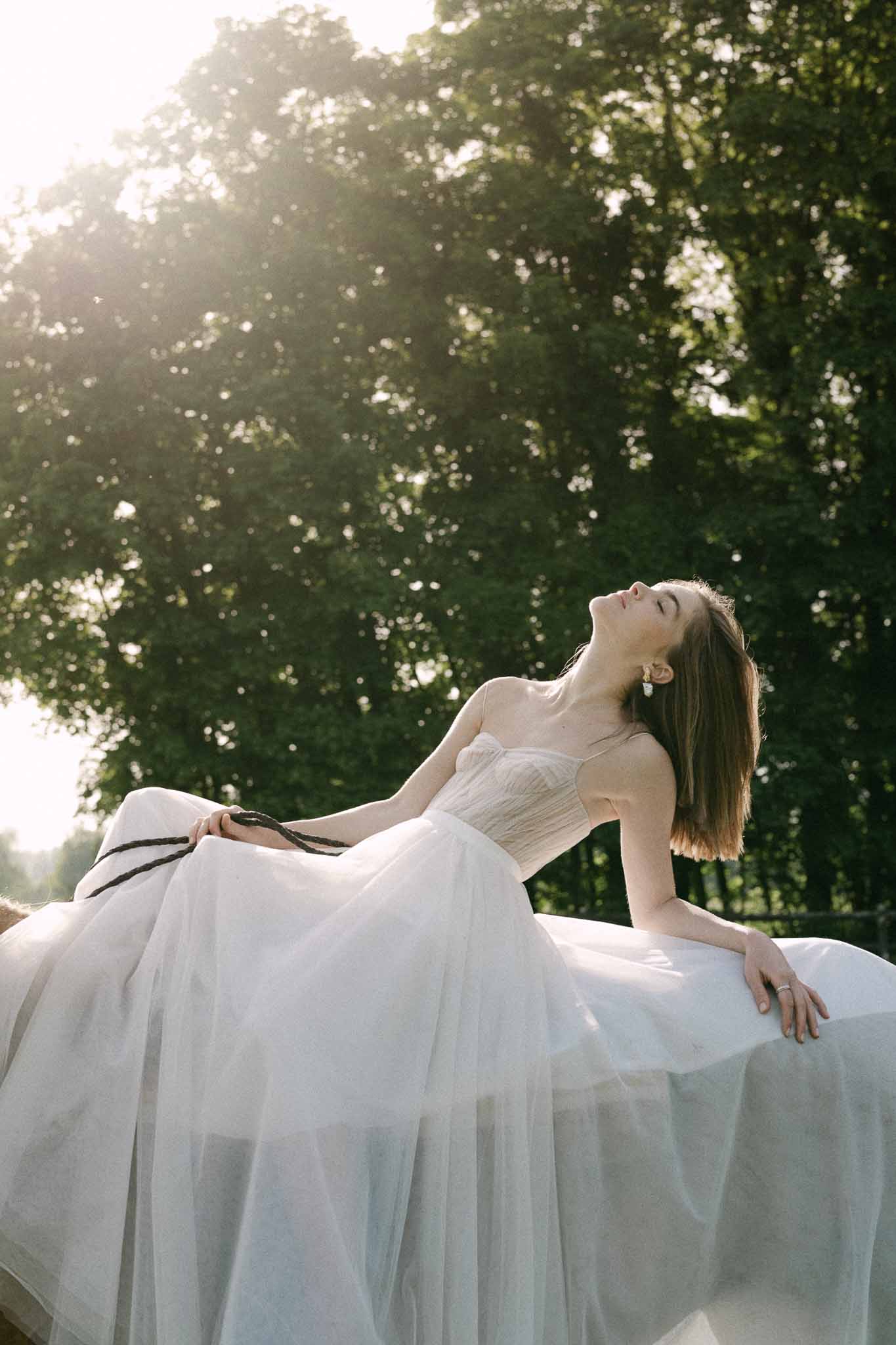 Bride in ivory strapless gown seated in outdoor garden with dappled sunlight filtering through trees