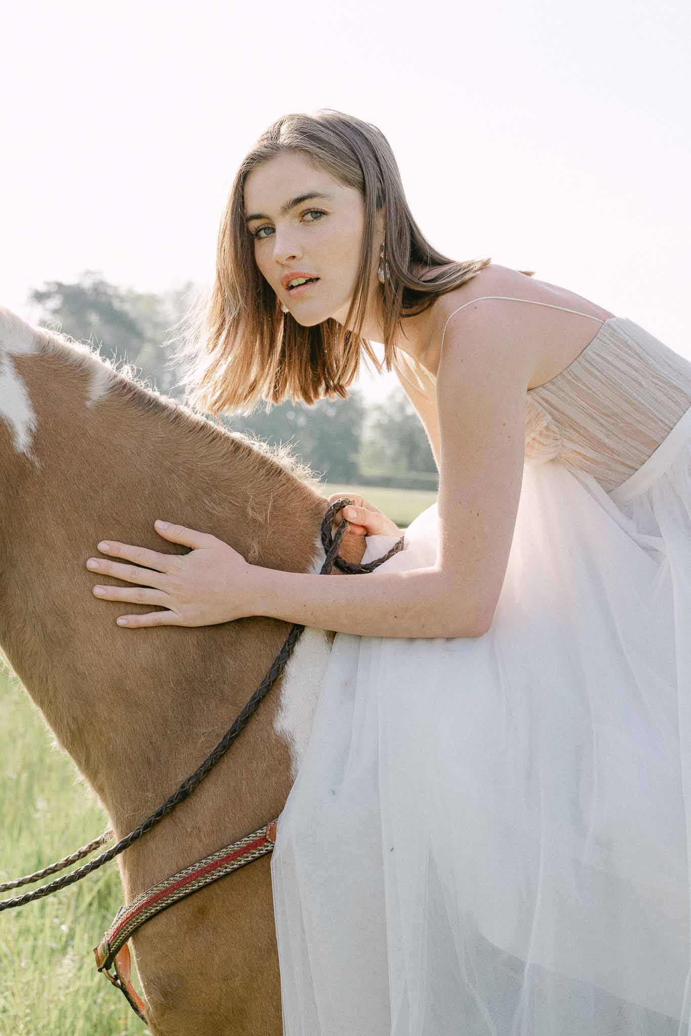 Bride in cream tulle gown posing with brown horse in countryside field