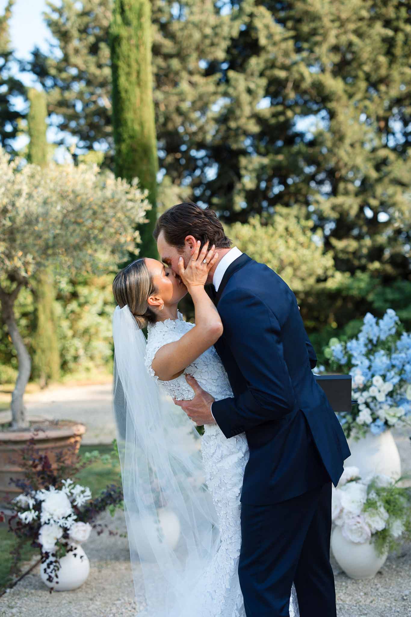 Bride and groom first kiss during garden ceremony with cypress trees and white florals