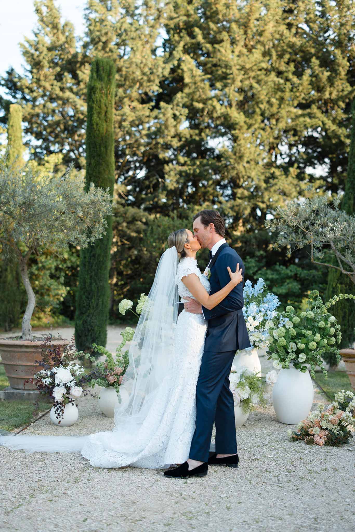 Bride and groom kissing in garden setting with cypress trees and white hydrangeas