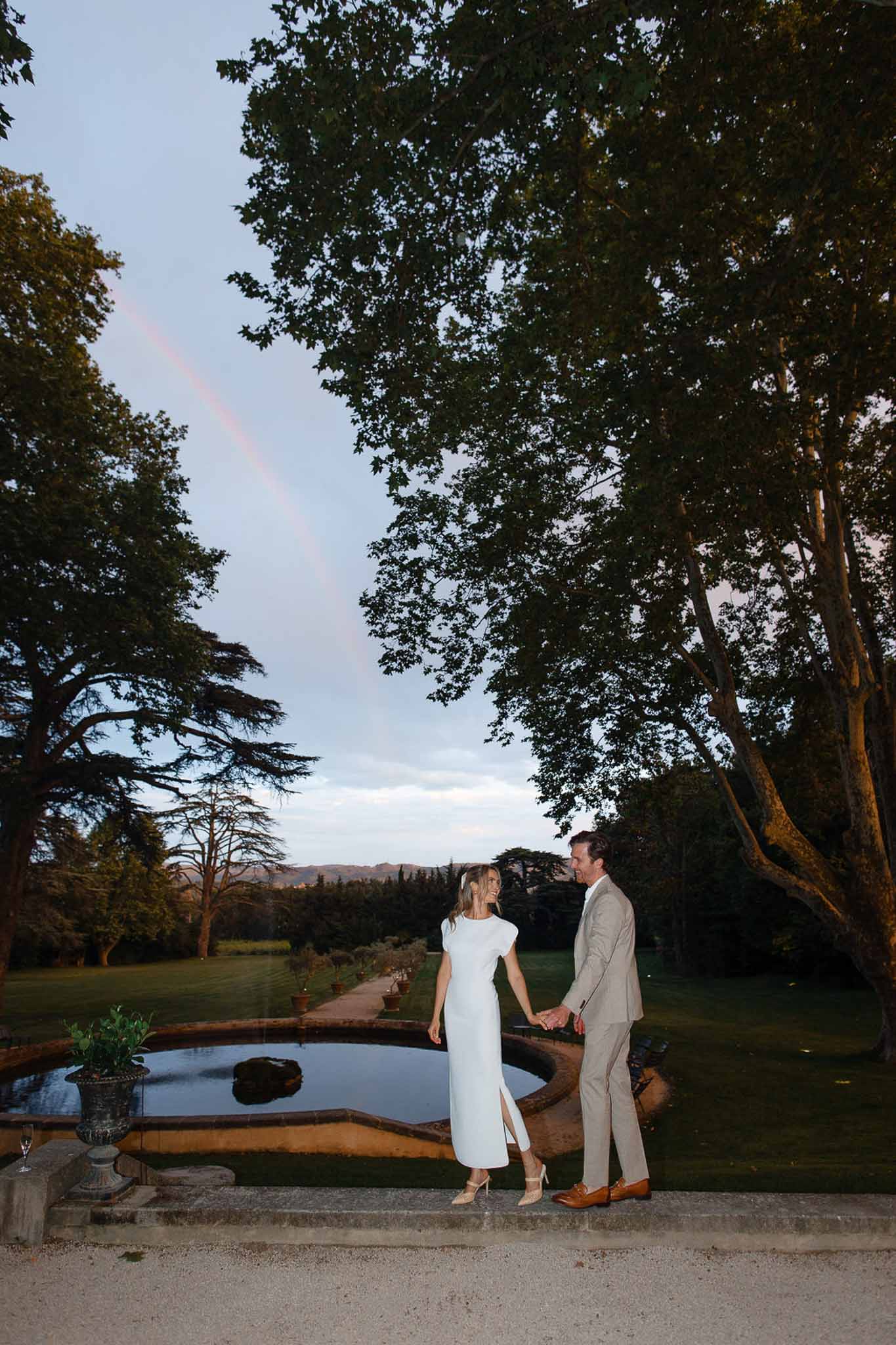 Bride and groom portrait beside stone pond in expansive garden with mature trees and distant hills