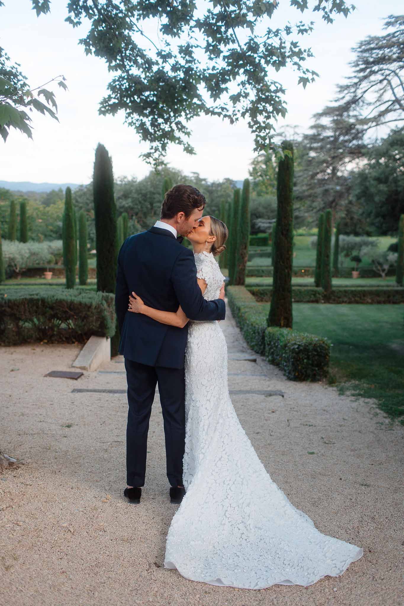 Bride and groom kissing on cypress-lined pathway in formal garden setting