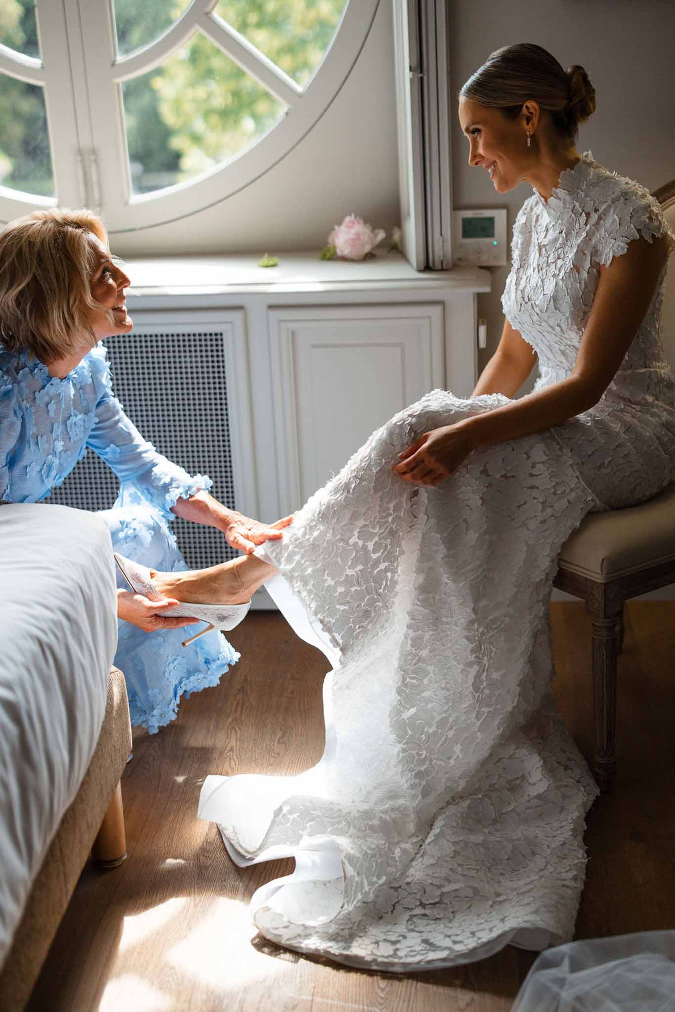Bride in lace wedding dress being attended to by mother during getting ready preparations indoors