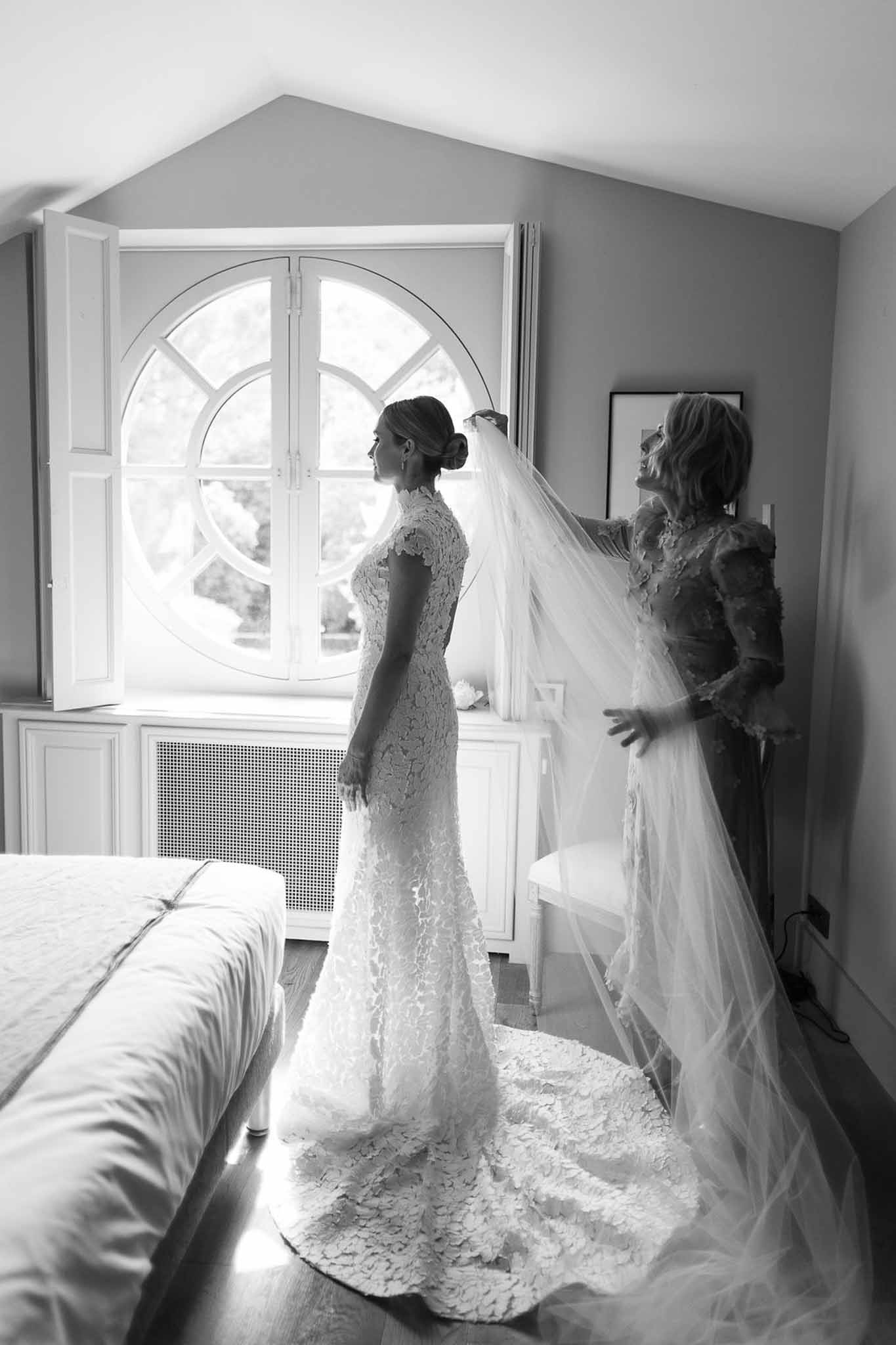 Bride having veil adjusted by helper in elegant bedroom with natural light