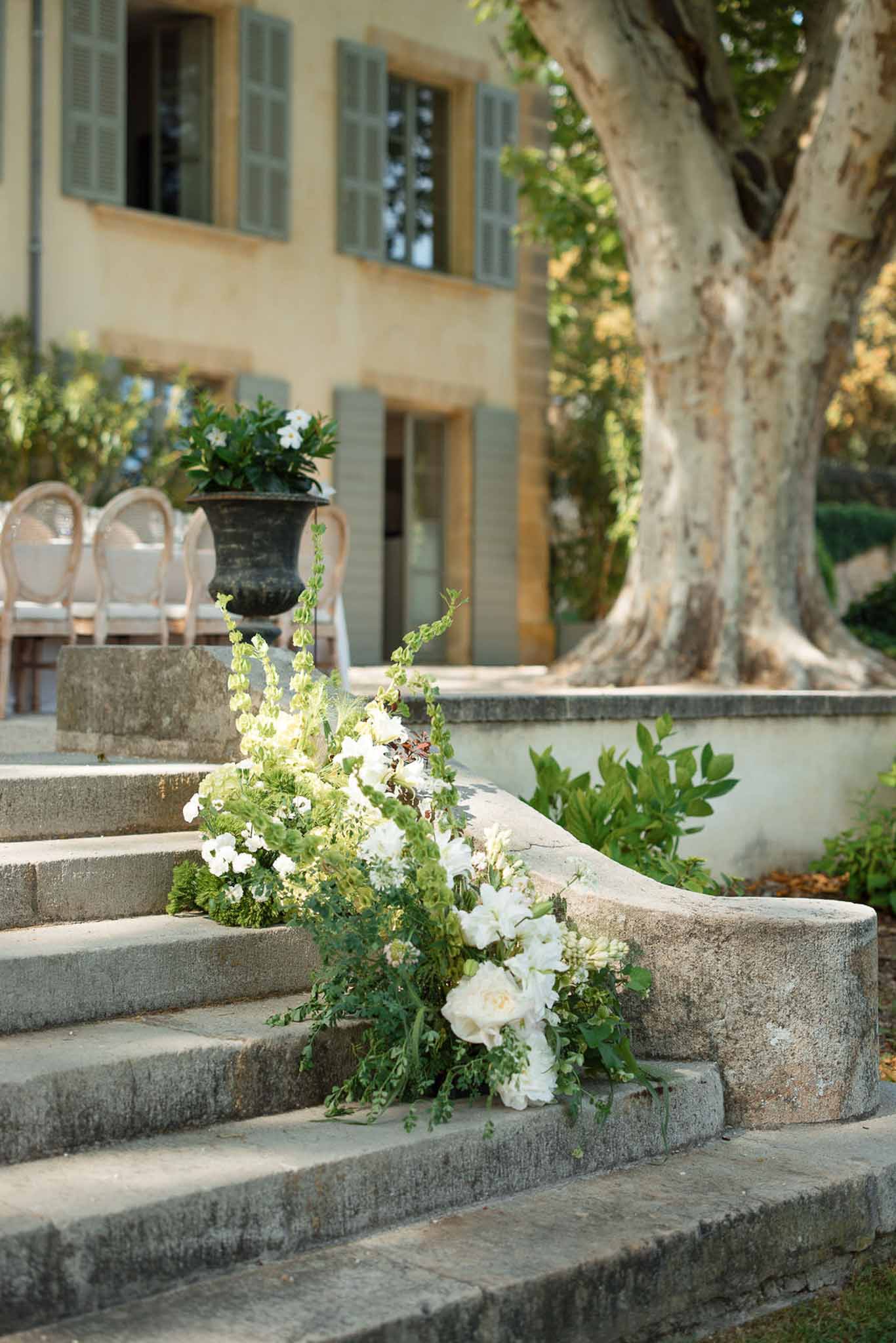 White floral installations on stone steps in Mediterranean courtyard with cream building and green shutters